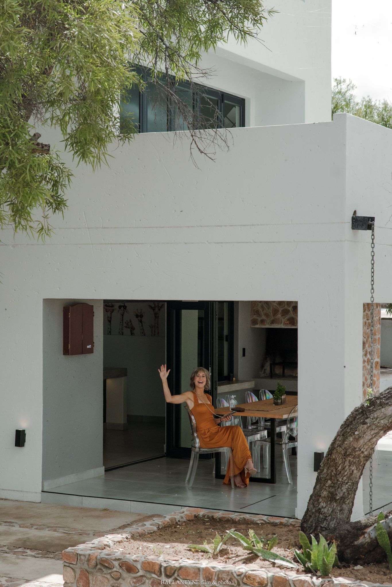 Woman in orange dress waving from open-plan kitchen of modern white Mediterranean villa with stone accents.