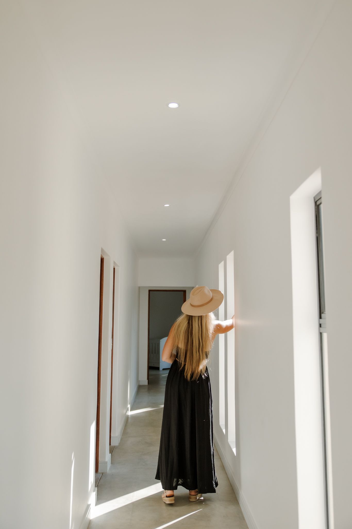 Someone in a long black dress and straw hat stands in a bright white hallway with natural light streaming through windows.