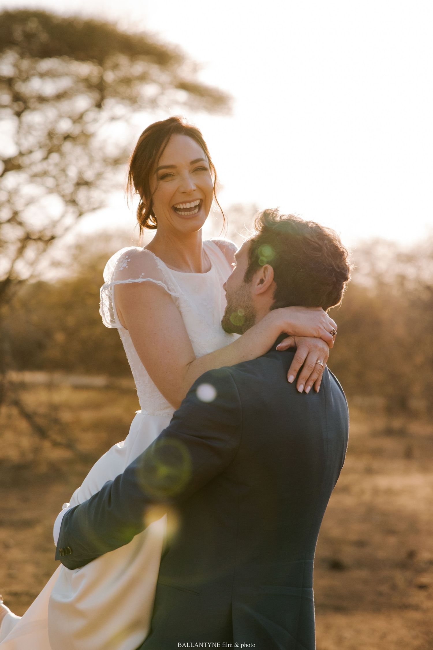 A bride in white dress laughs while being lifted by her partner during golden hour.