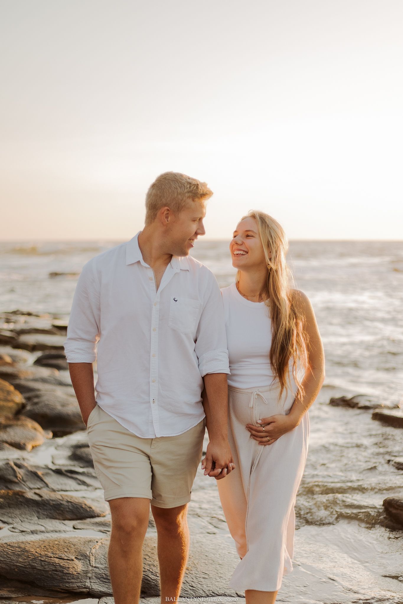 Couple in white outfits walking along rocky beach at sunset.