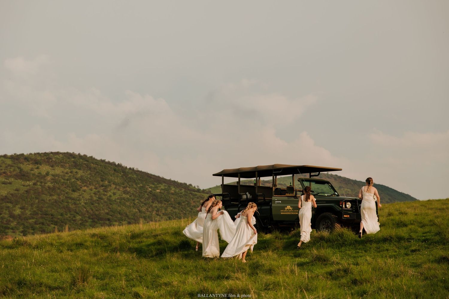 Wedding party in white dresses gathered around a safari vehicle on a grassy hillside.