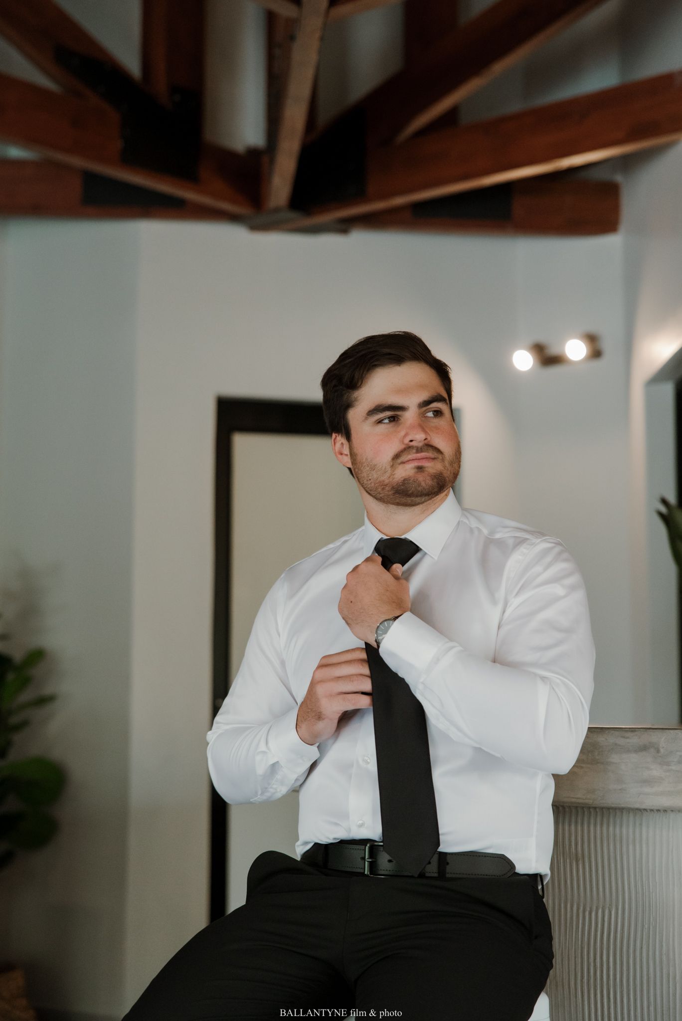 Groom in white dress shirt adjusting black tie while seated, preparing for wedding ceremony indoors.