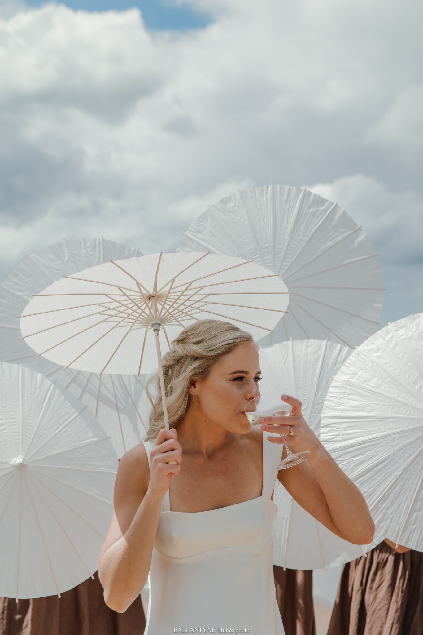 Bride in white dress holding parasol surrounded by white umbrellas against cloudy sky.