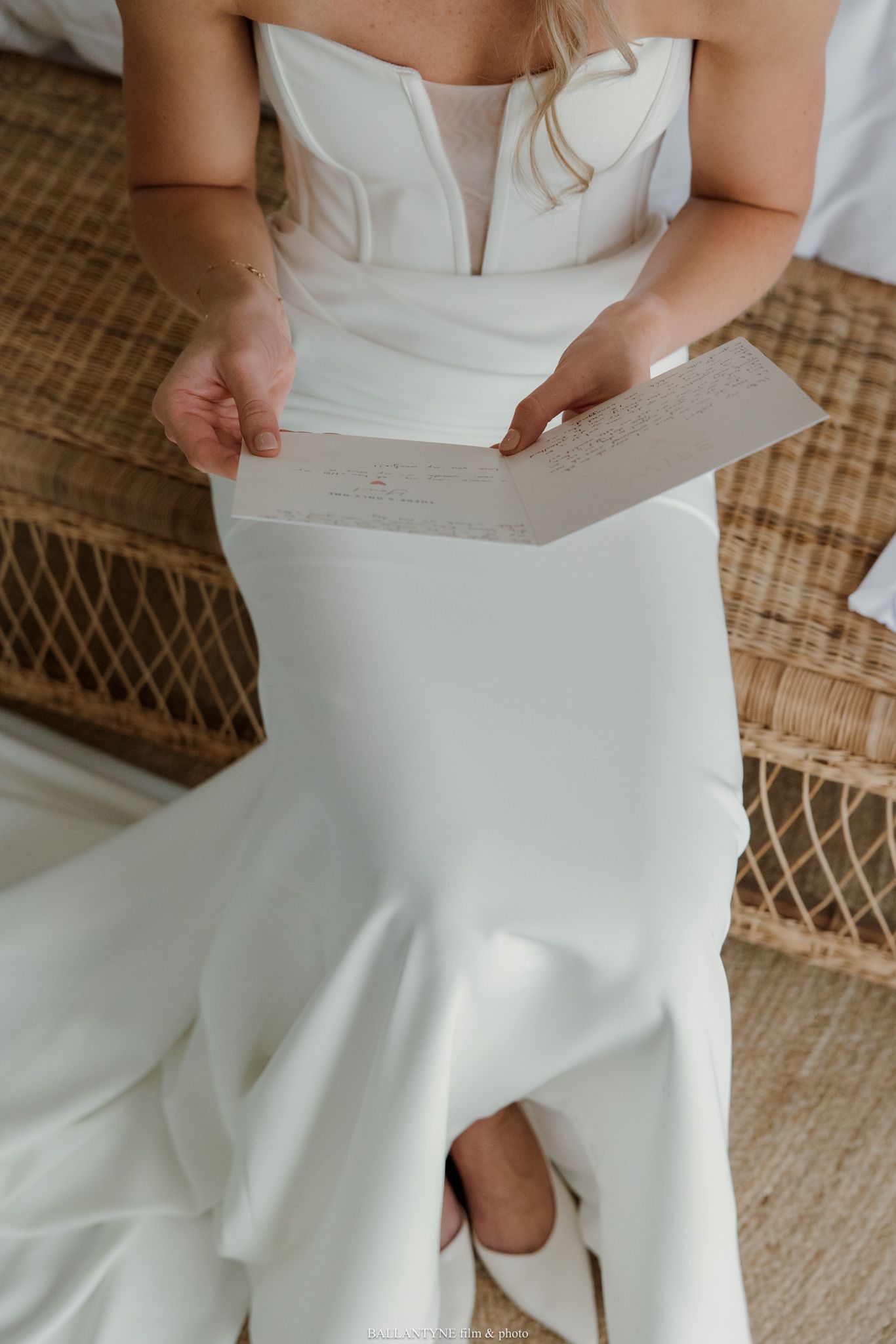 Bride in strapless white mermaid gown reading a letter while seated on a wicker chair on her wedding day.