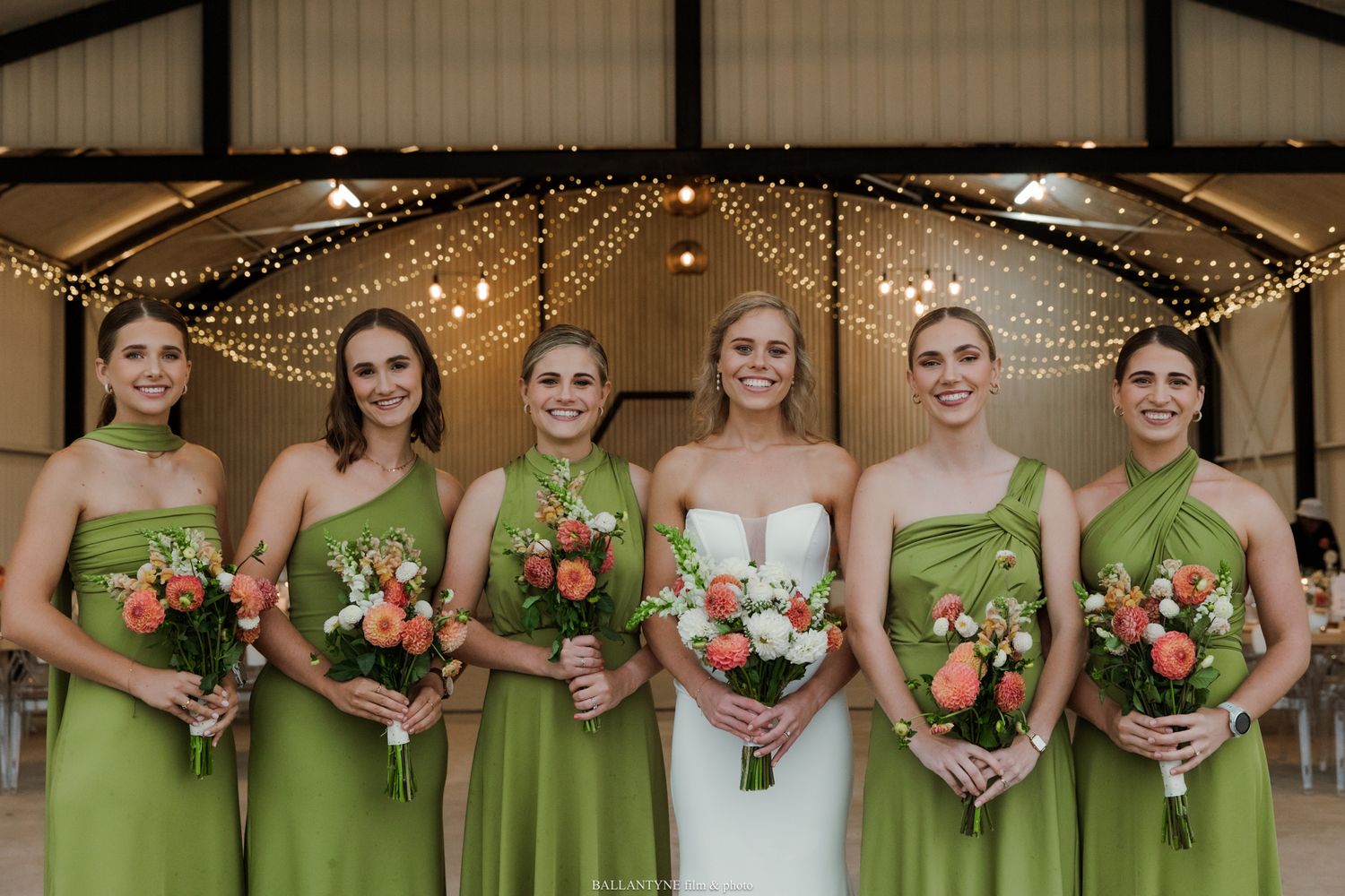Bride in white gown with five bridesmaids in green dresses holding coral bouquets at elegant barn wedding venue.