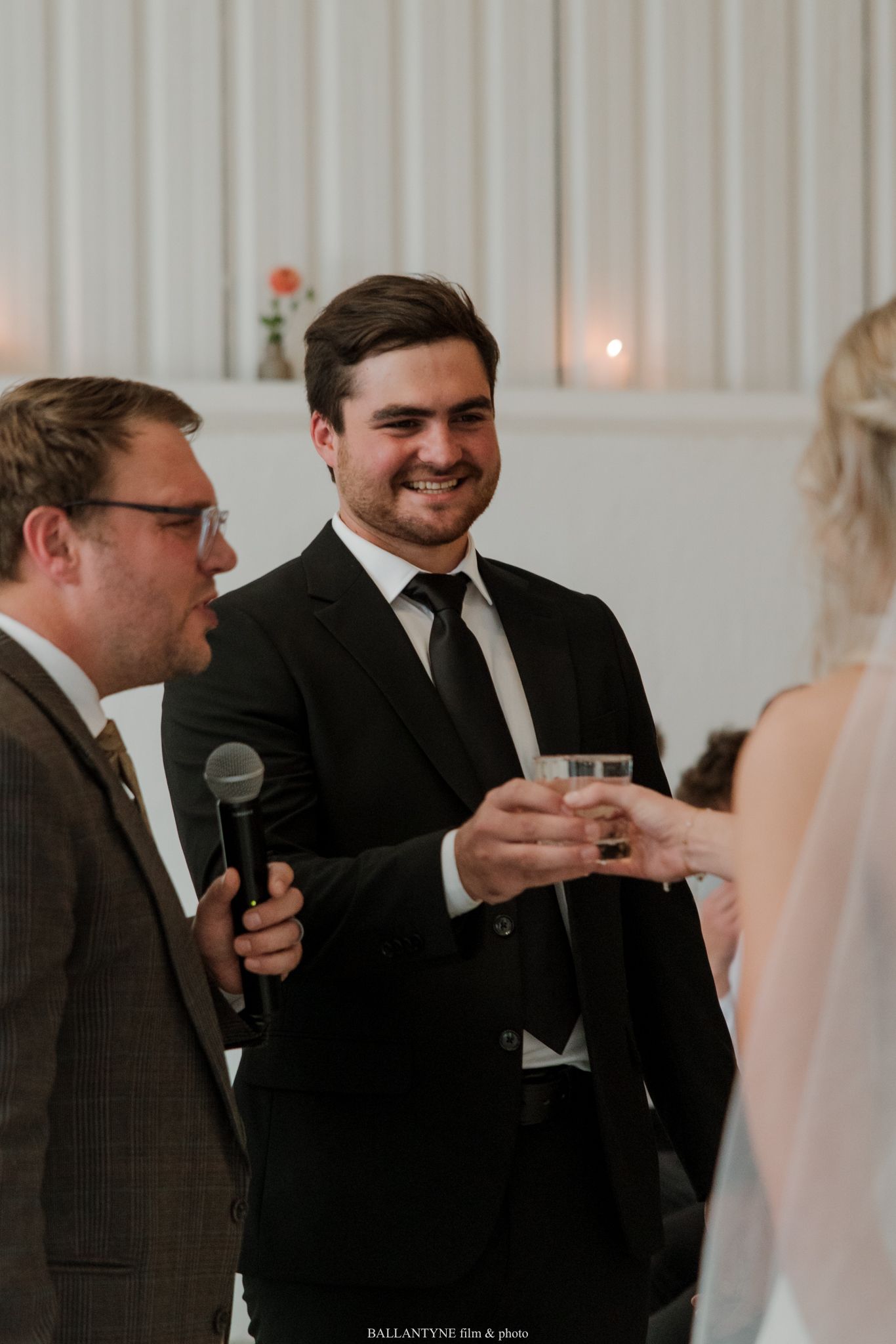Groom in black suit smiling and raising a glass during wedding reception toast with officiant holding microphone nearby.