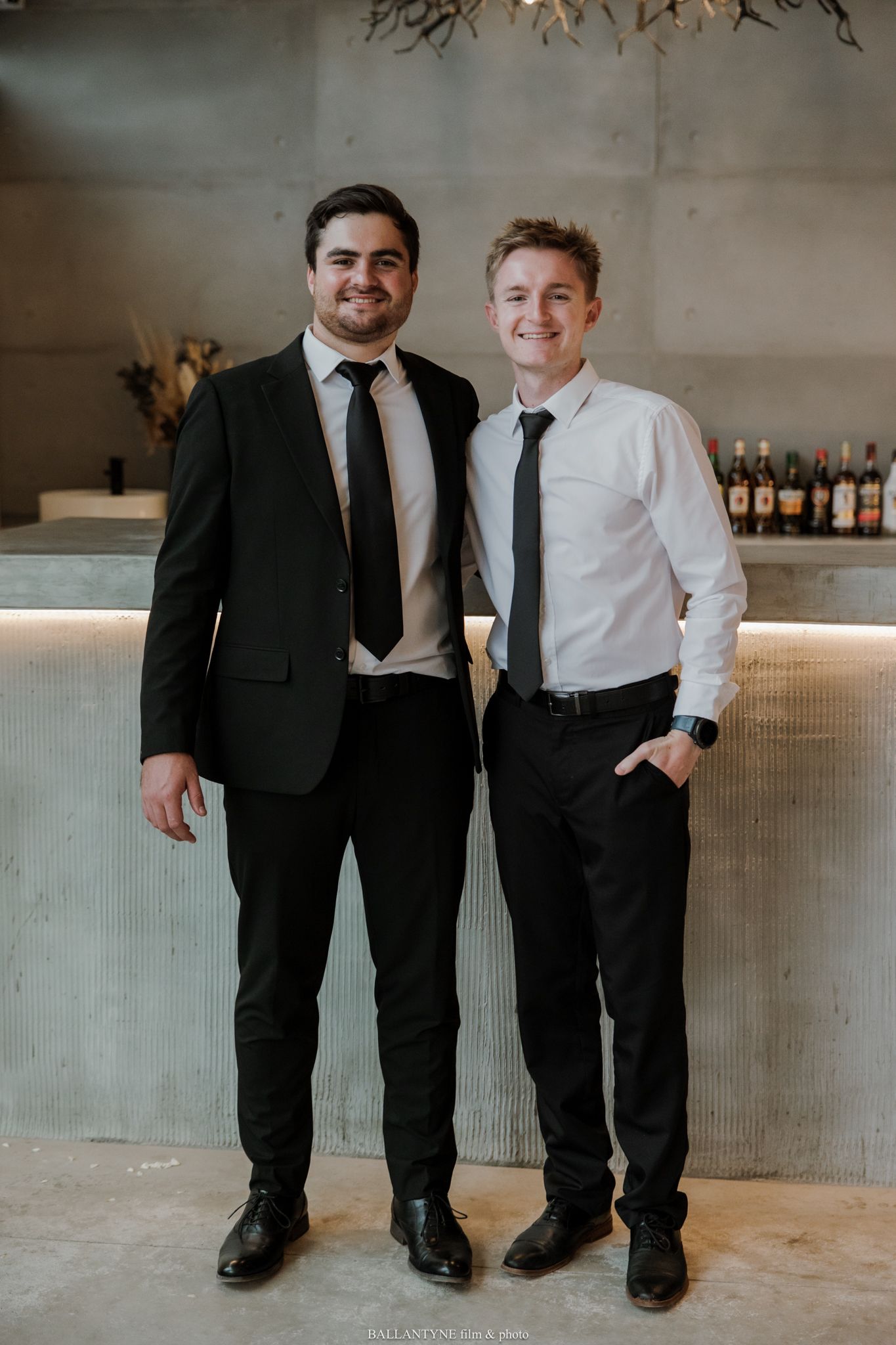Two well-dressed men in black ties and dress shirts smile together in front of a modern bar backdrop.