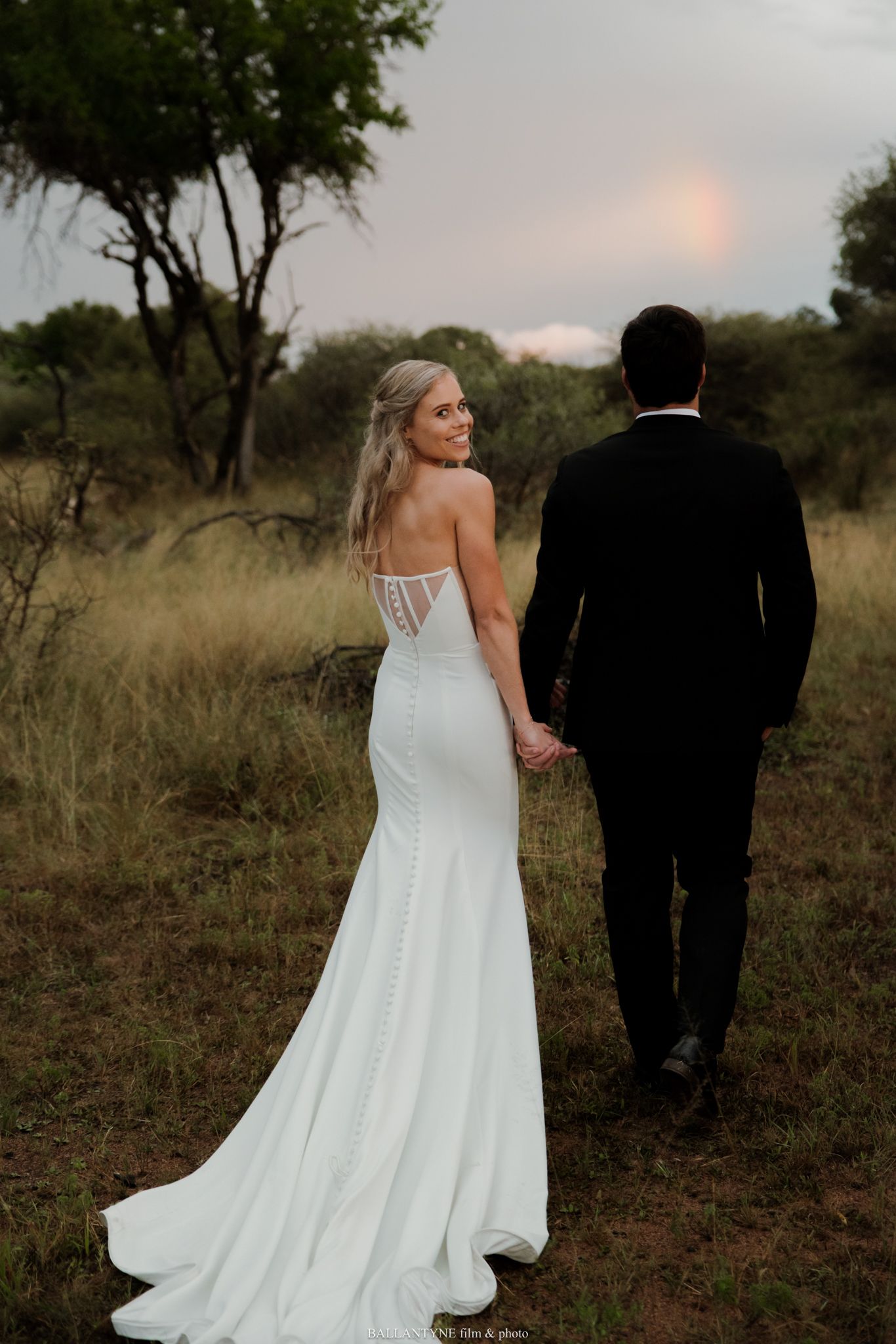 Bride in elegant backless gown and groom in black tuxedo walk hand in hand at dusk.