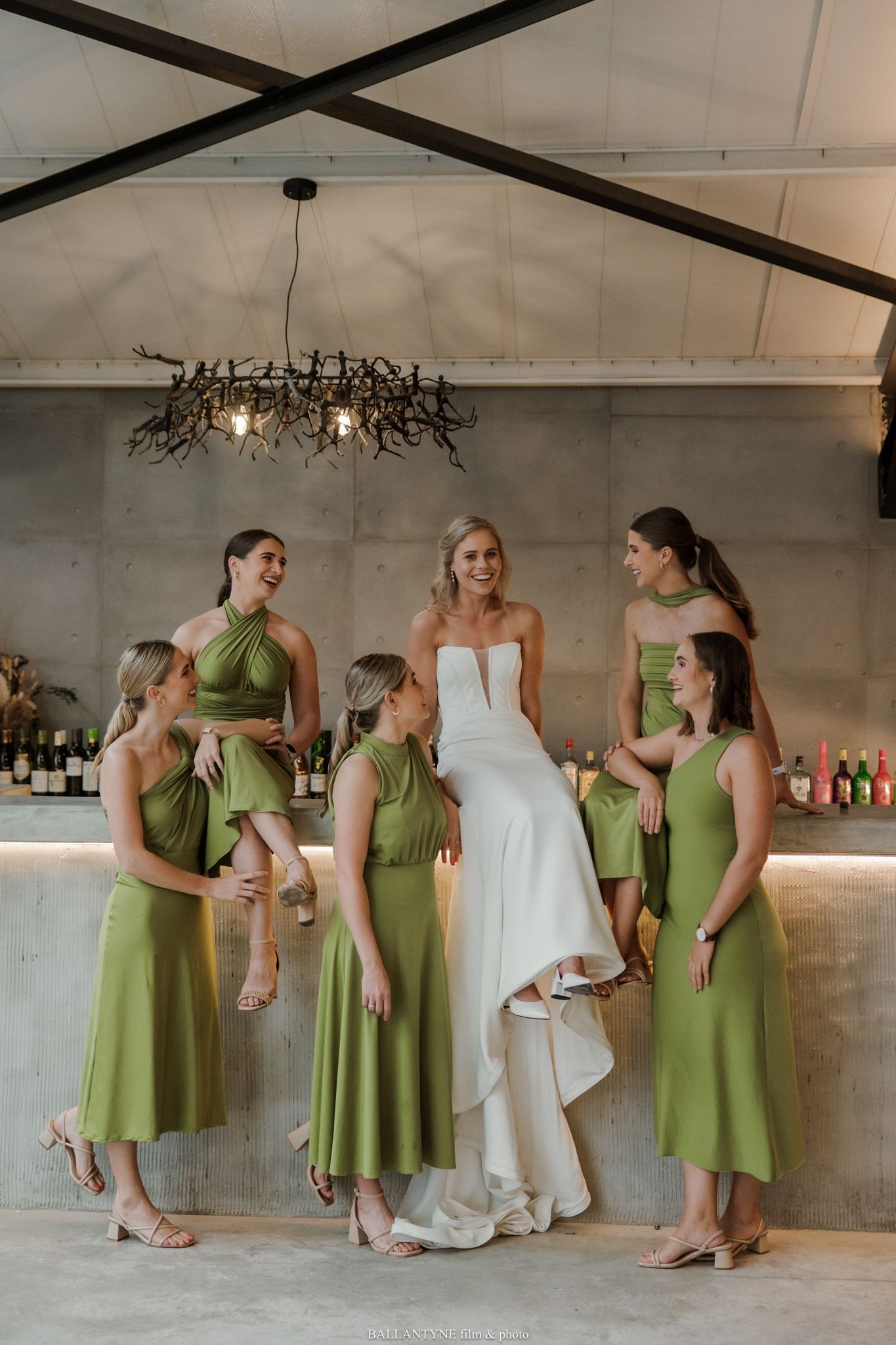 Bride in white gown with four bridesmaids in olive green dresses posing at a modern wedding venue bar.