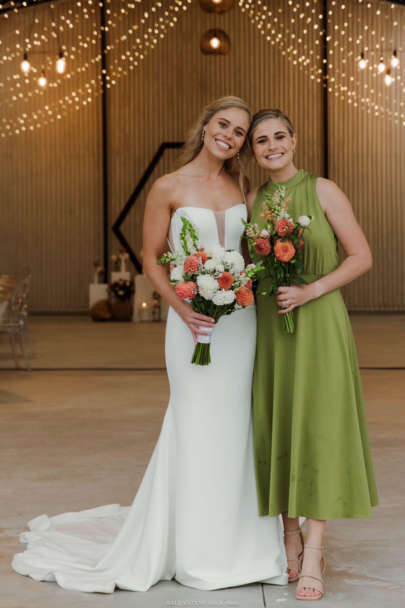 Bride in white gown and bridesmaid in green dress holding coral and white bouquets at a wedding venue.