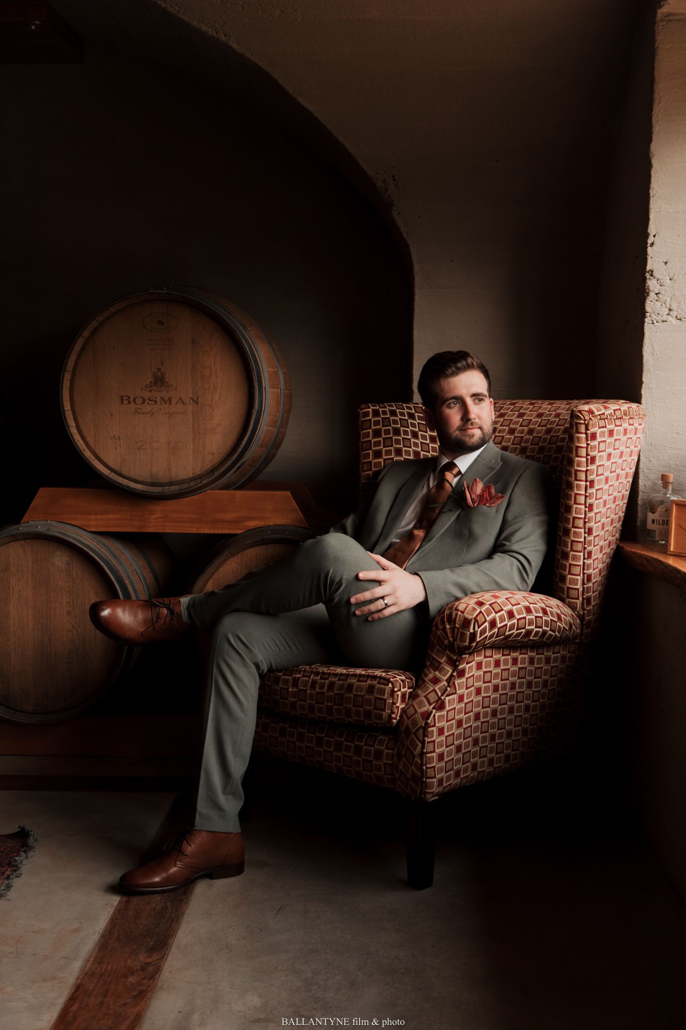 Person in olive suit relaxes on vintage leather couch in moody wine cellar setting.
