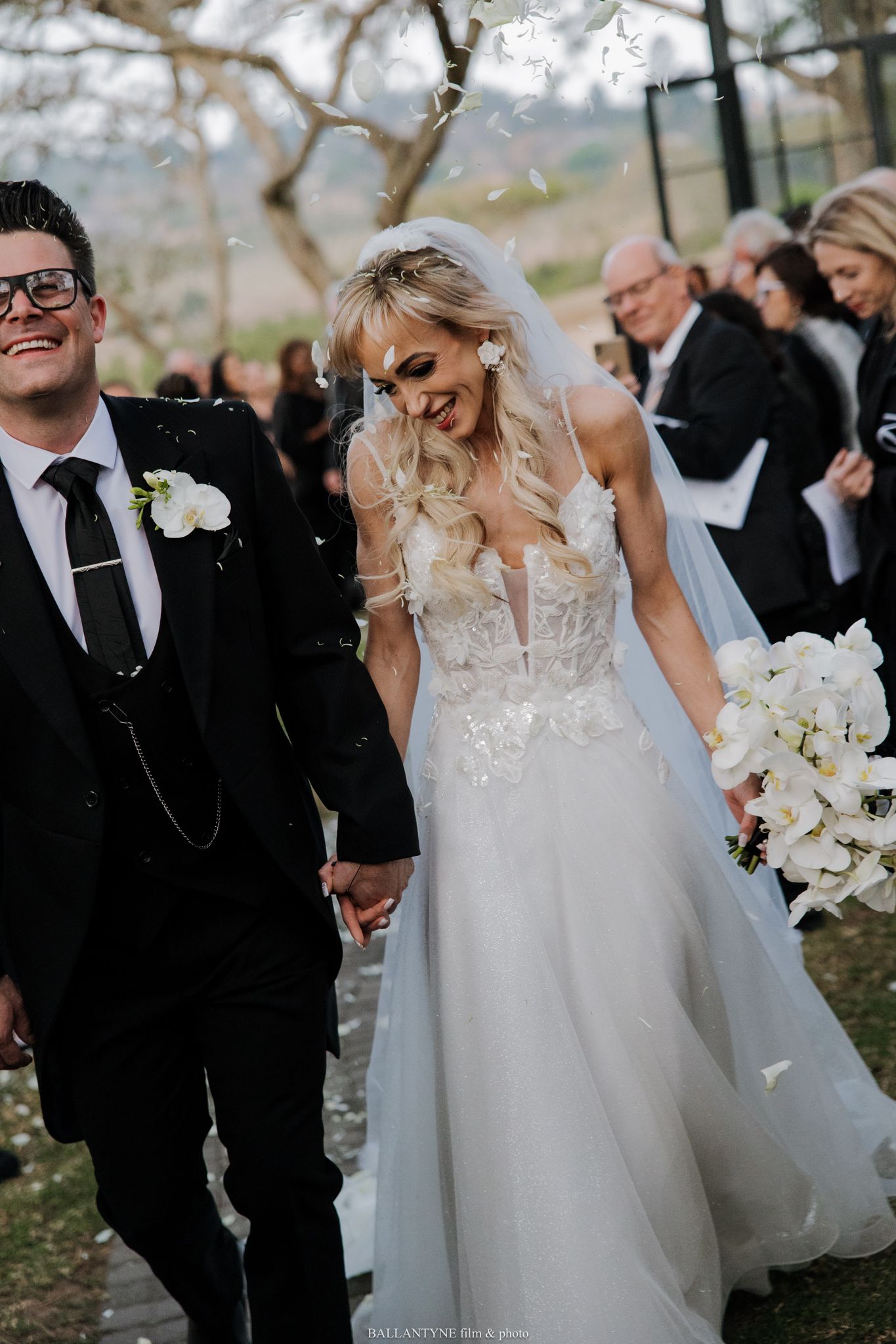 Wedding couple walking outdoors with guests in background.