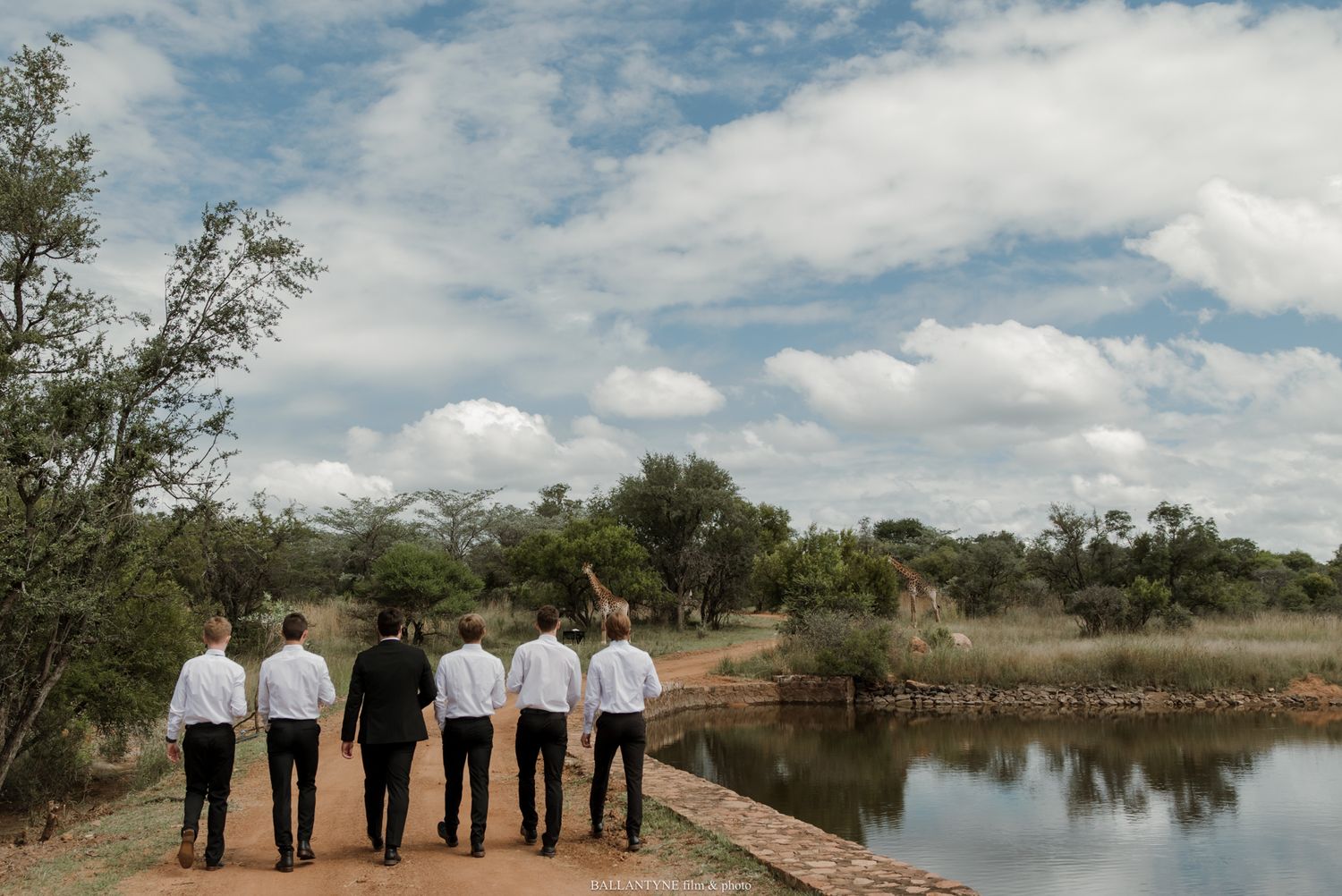 Six groomsmen in white shirts walk along a dirt path beside a pond in a scenic African bush landscape.