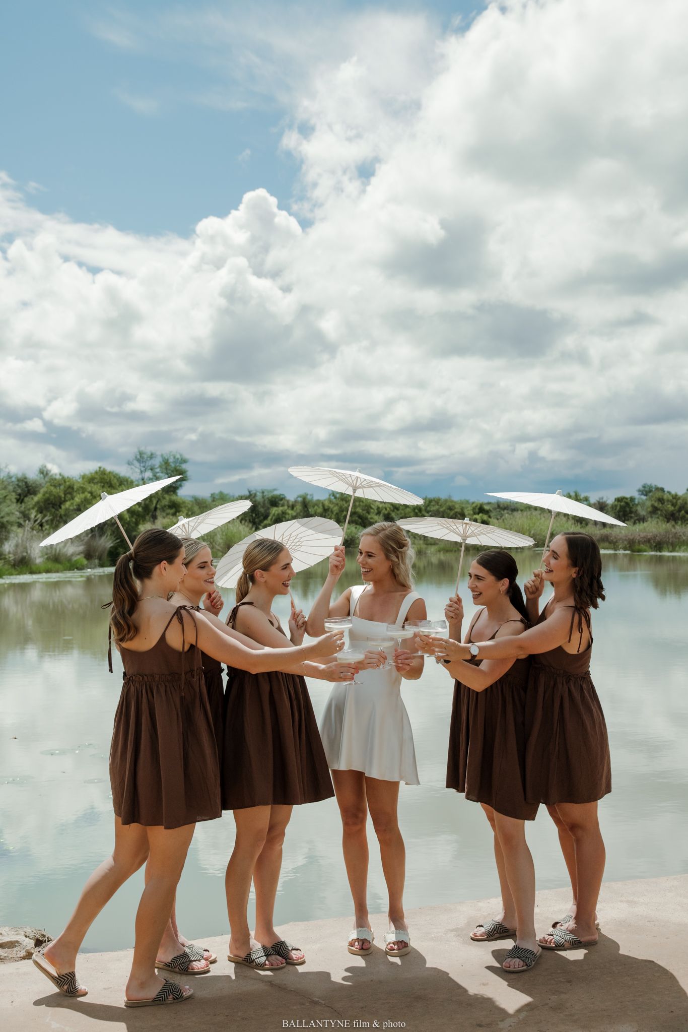 Bride in white dress with bridesmaids in brown dresses holding white parasols by a lake under a cloudy sky.