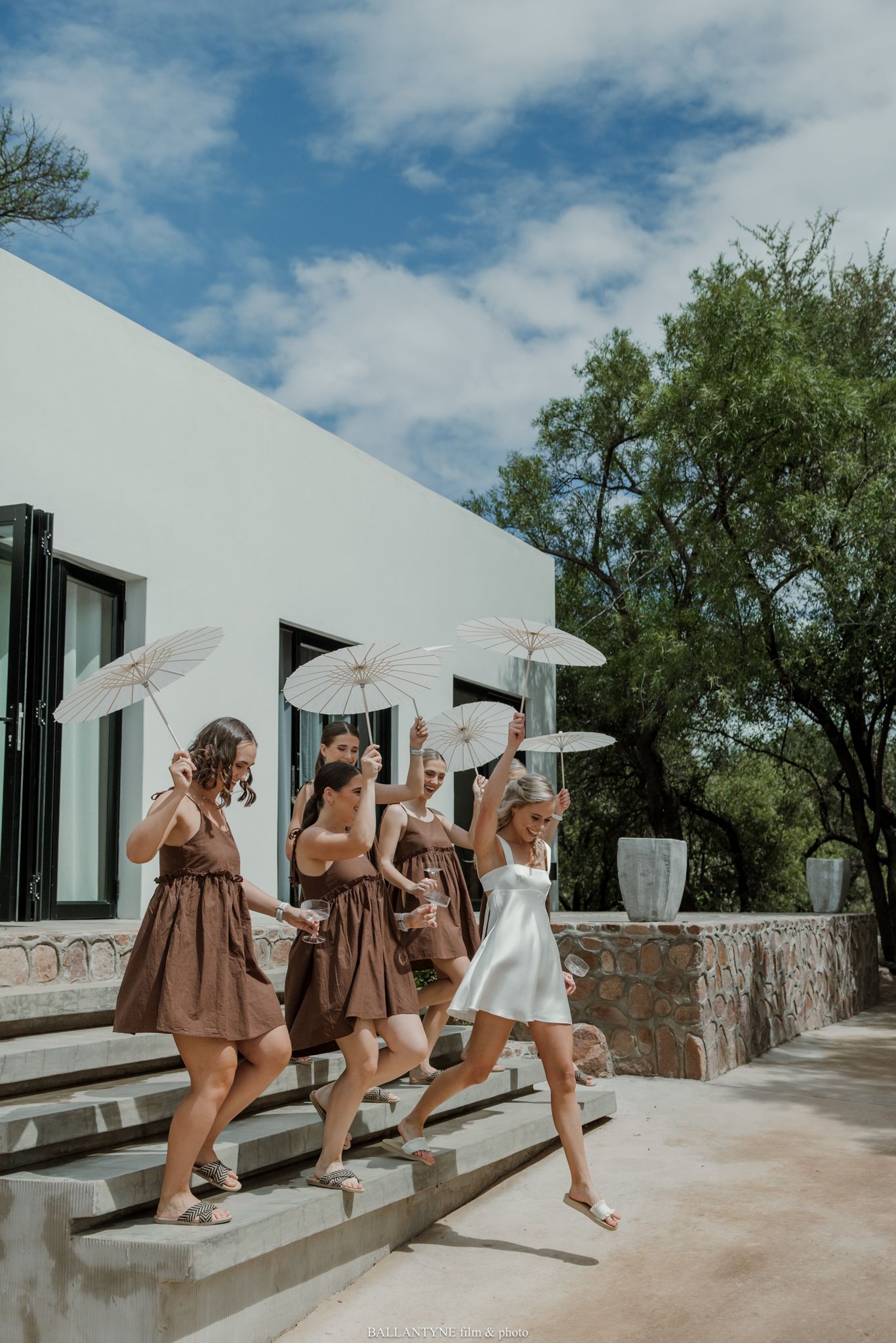 Bride in white dress leads bridesmaids in brown dresses down steps holding white parasols at outdoor wedding venue.