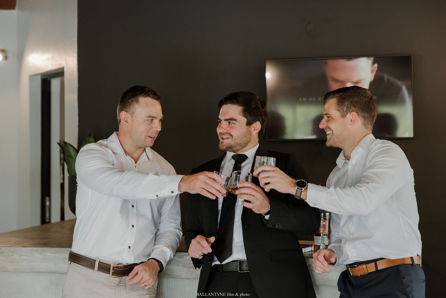 Three groomsmen in formal attire toasting with drinks during a wedding celebration indoors.