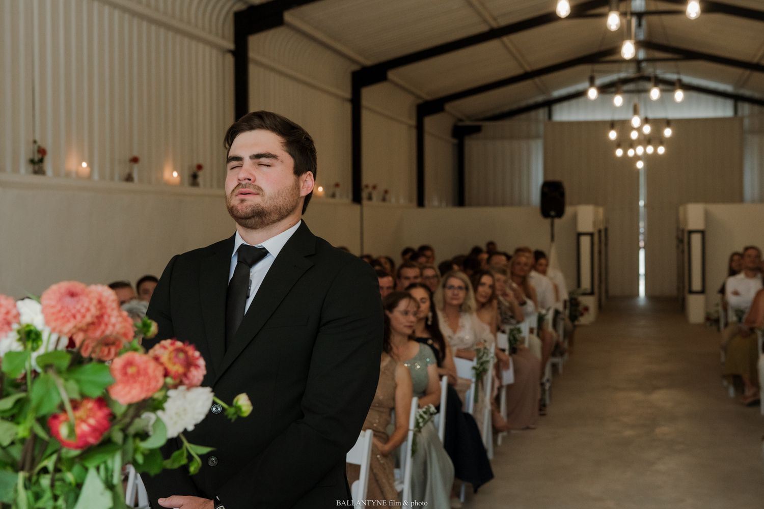 Groom in black suit stands at wedding altar in decorated barn venue, awaiting bride as guests fill white chairs.