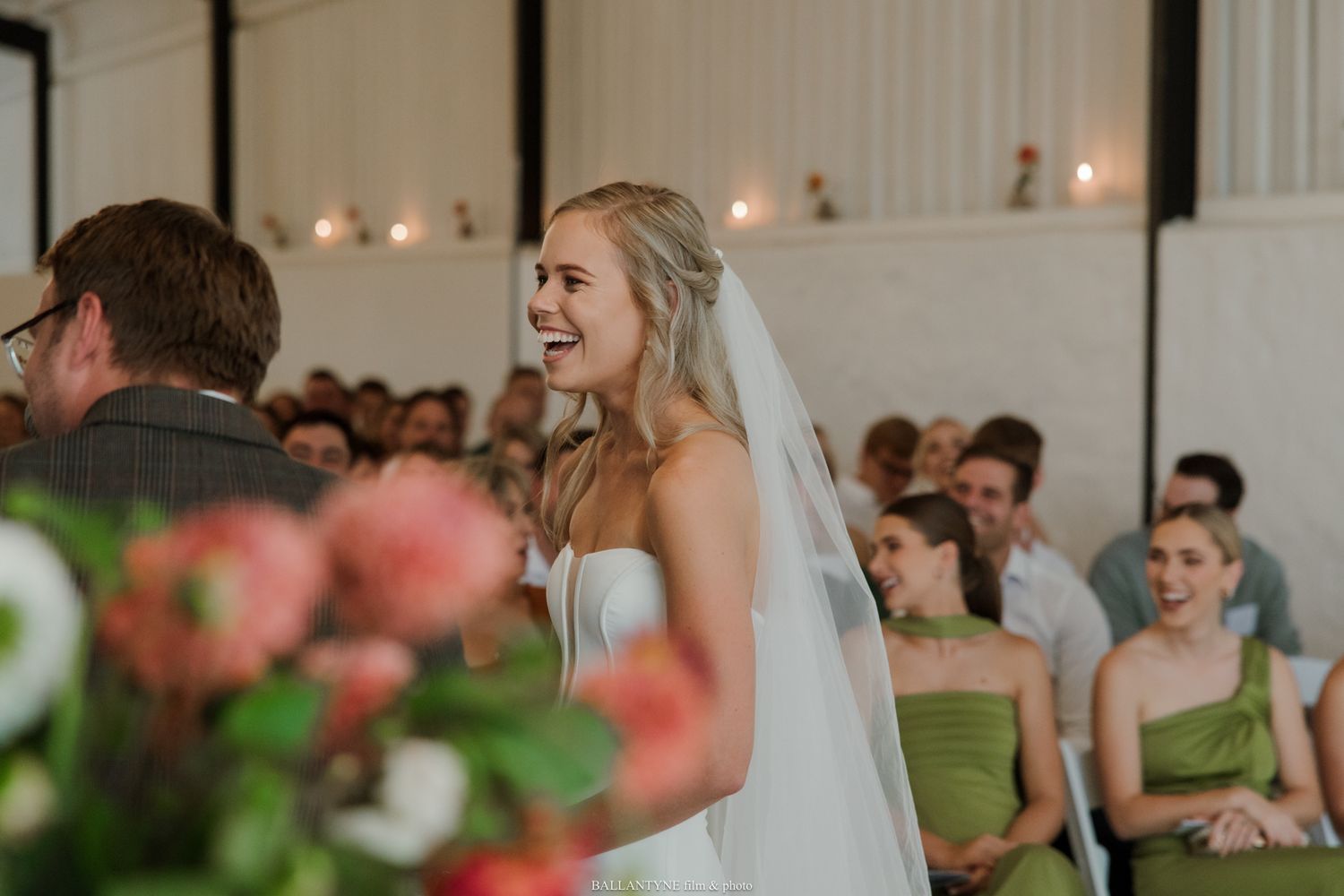 Bride laughing joyfully at wedding ceremony altar, wearing strapless gown and veil, with guests and bridesmaids in green dresses.