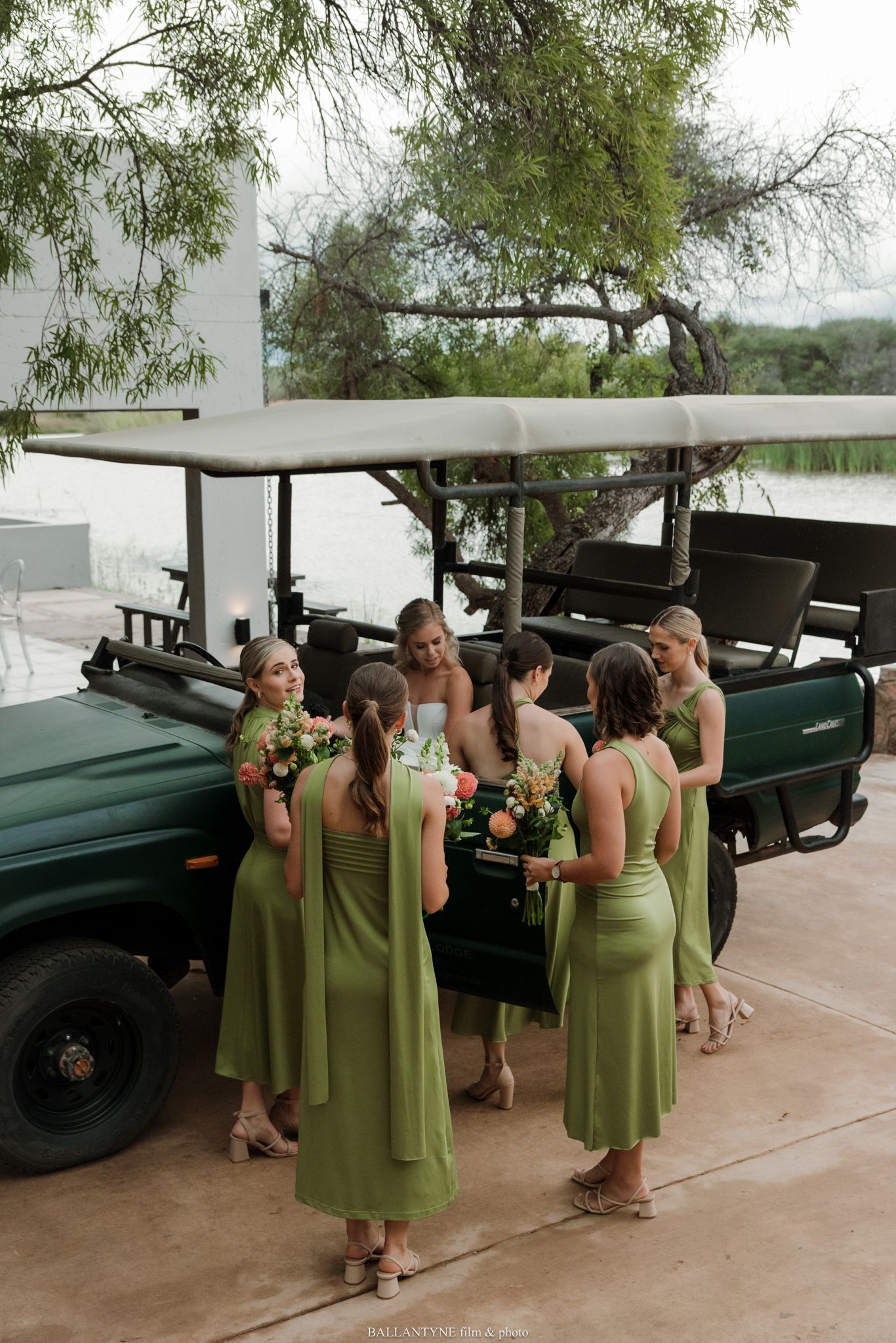 Bridesmaids in olive green dresses holding bouquets gather beside a dark green safari truck at an outdoor wedding.