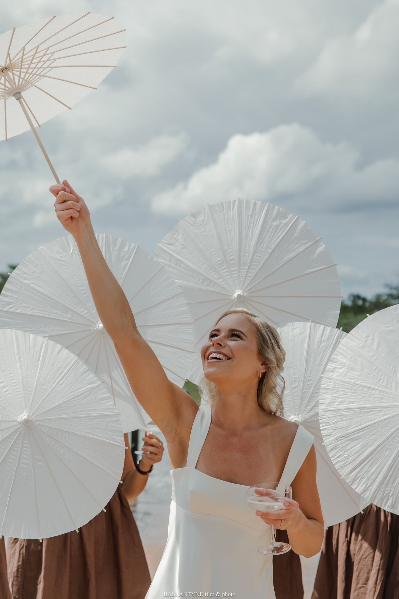 Smiling bride in white dress holding parasol aloft among white paper umbrellas at outdoor wedding celebration.
