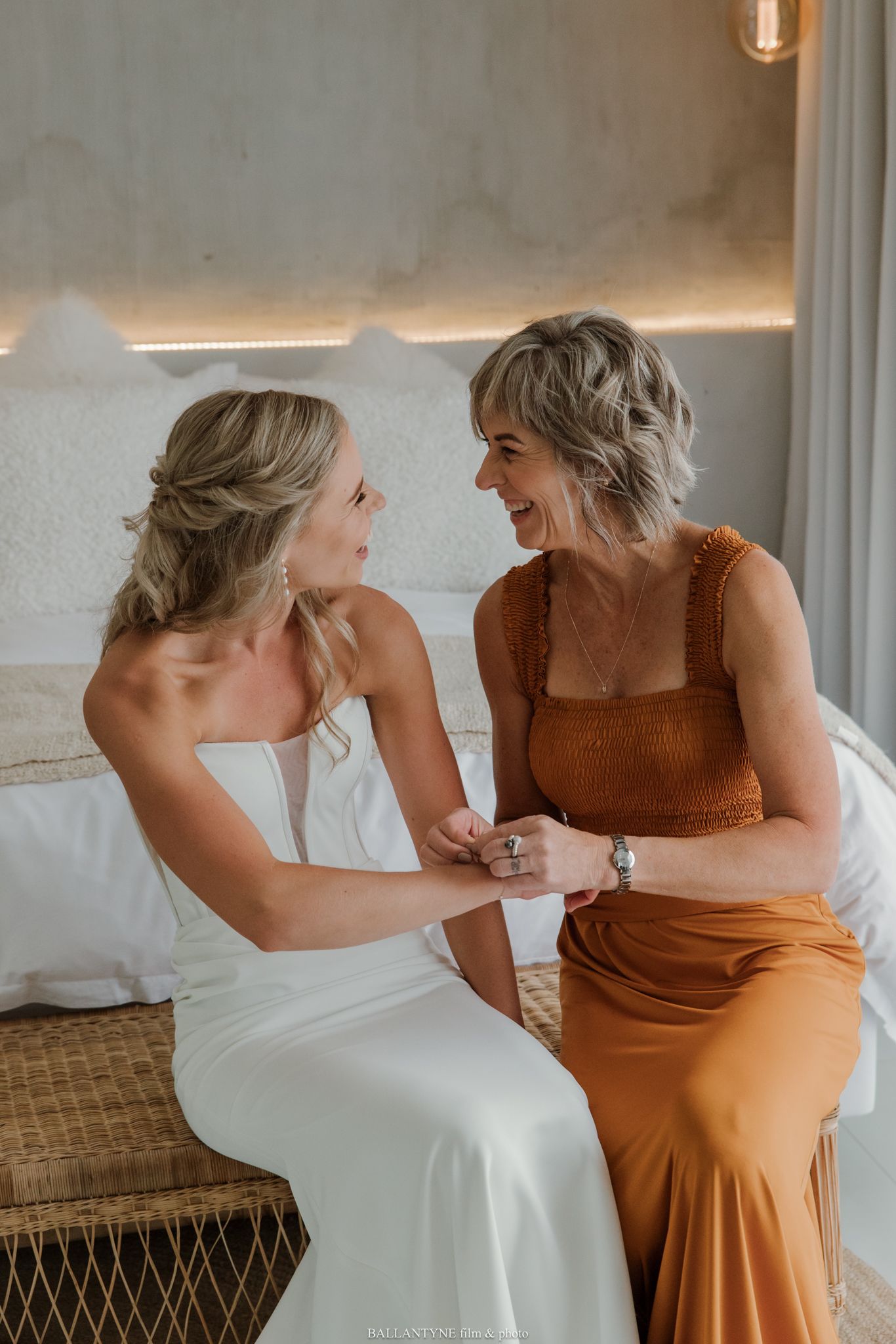 Bride in white gown and woman in rust orange dress sitting together smiling, holding hands on wedding day.