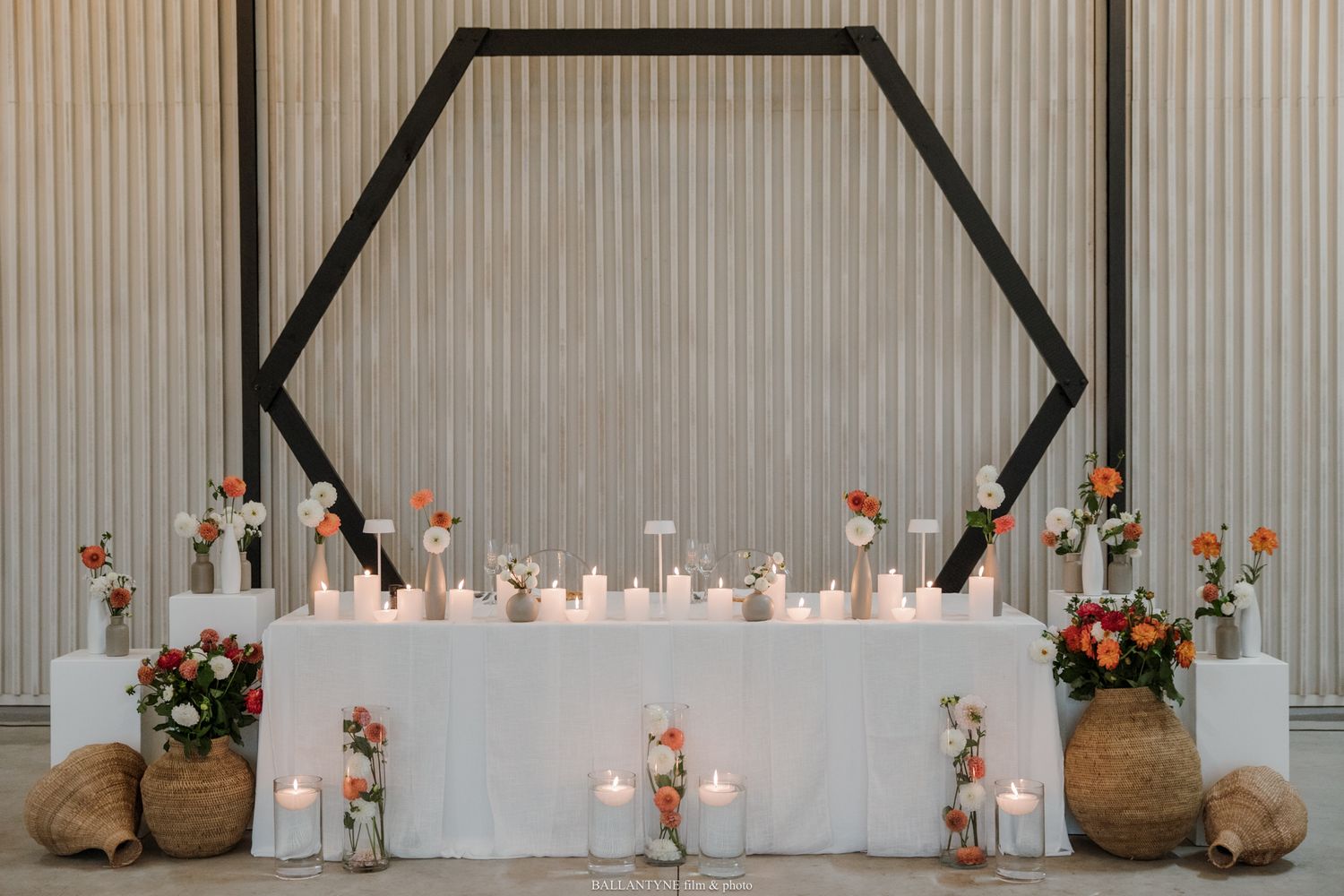 Elegant wedding sweetheart table with hexagon backdrop, white linens, candles, and coral floral arrangements.