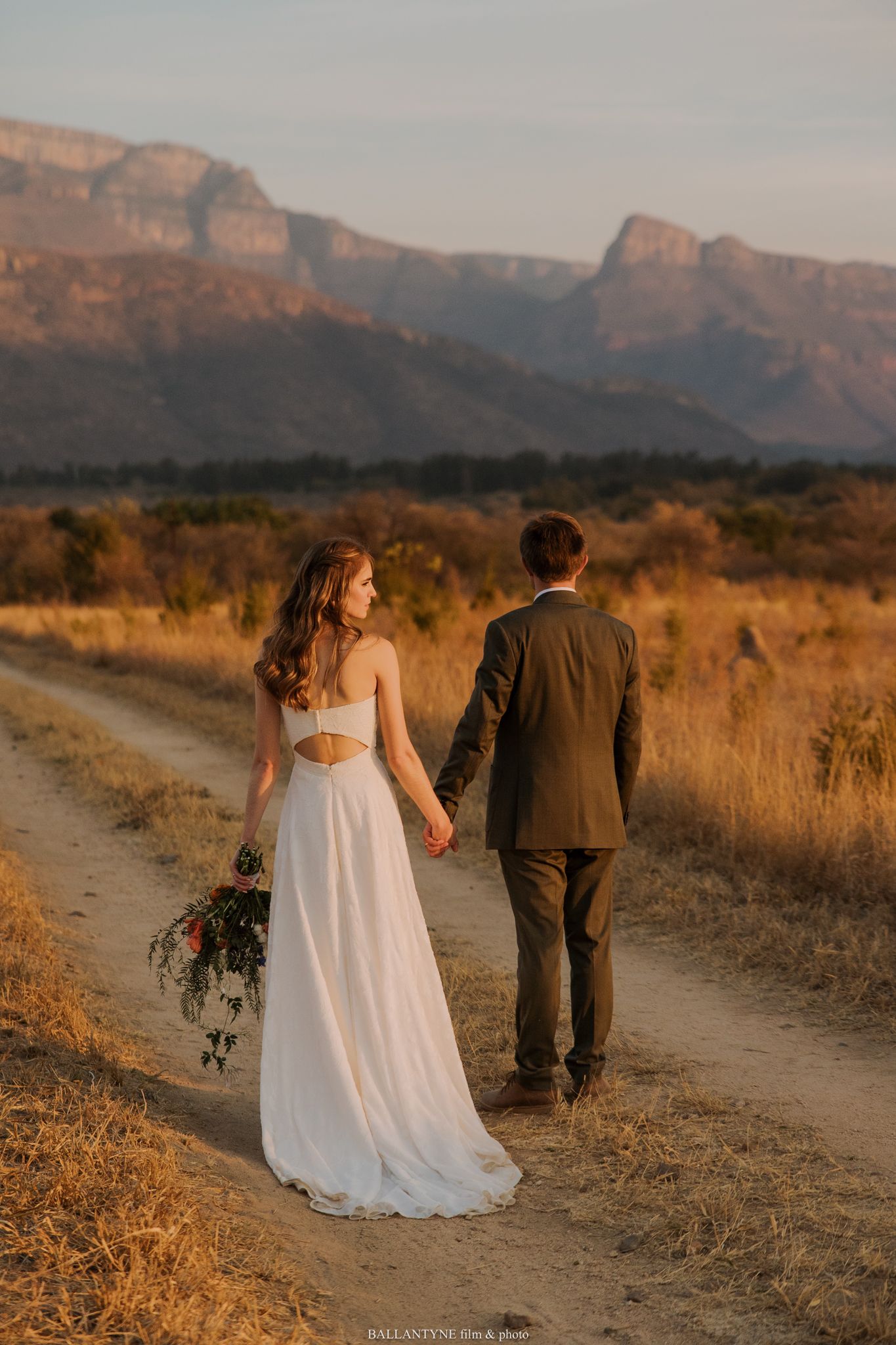 Couple walking hand in hand on dirt path with mountain landscape backdrop.