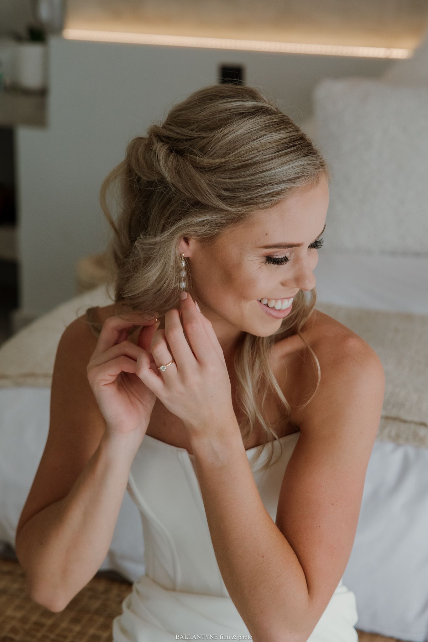 Smiling bride putting on pearl drop earrings while wearing a white dress, with an elegant updo hairstyle.