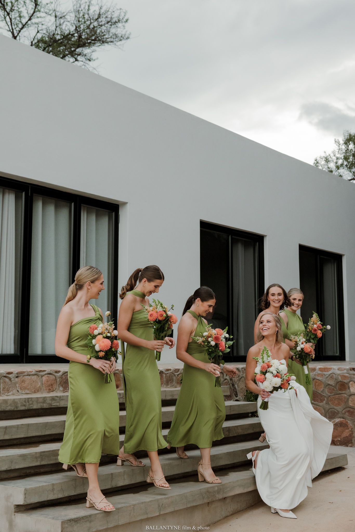 Bride in white gown poses on steps with bridesmaids in green dresses holding coral bouquets outside modern venue.