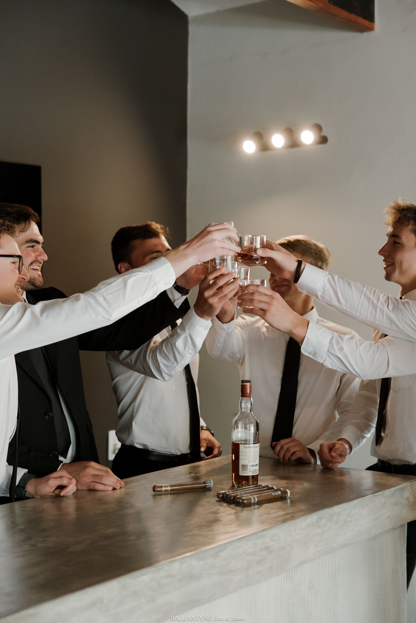 A group of groomsmen in white shirts and ties toasting with shot glasses over a whiskey bottle at a bar.