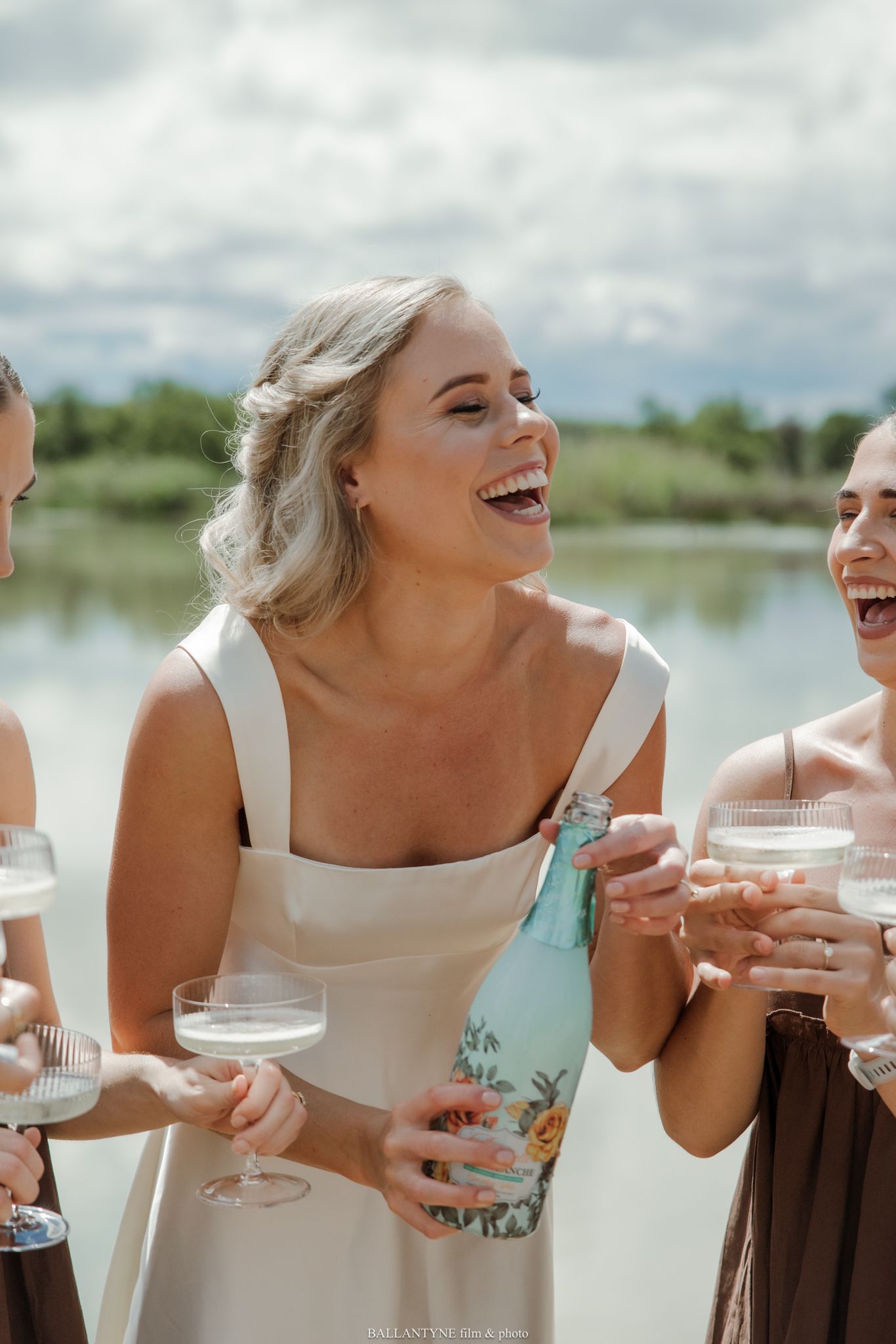 Laughing bride in white dress holding champagne bottle with bridesmaids in brown dresses near a lake.