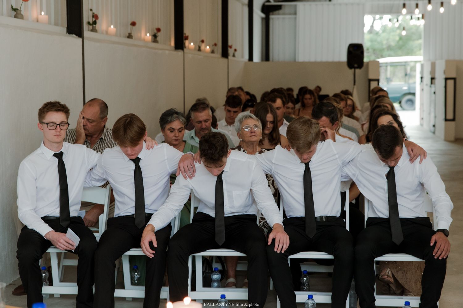 Young men in white shirts and black ties stand together in a row at a formal indoor wedding ceremony.