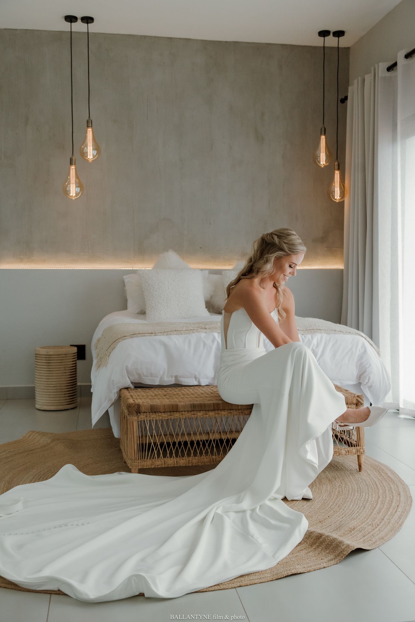 Bride in elegant white gown with long train sits on wicker bench in modern bedroom with pendant lights.