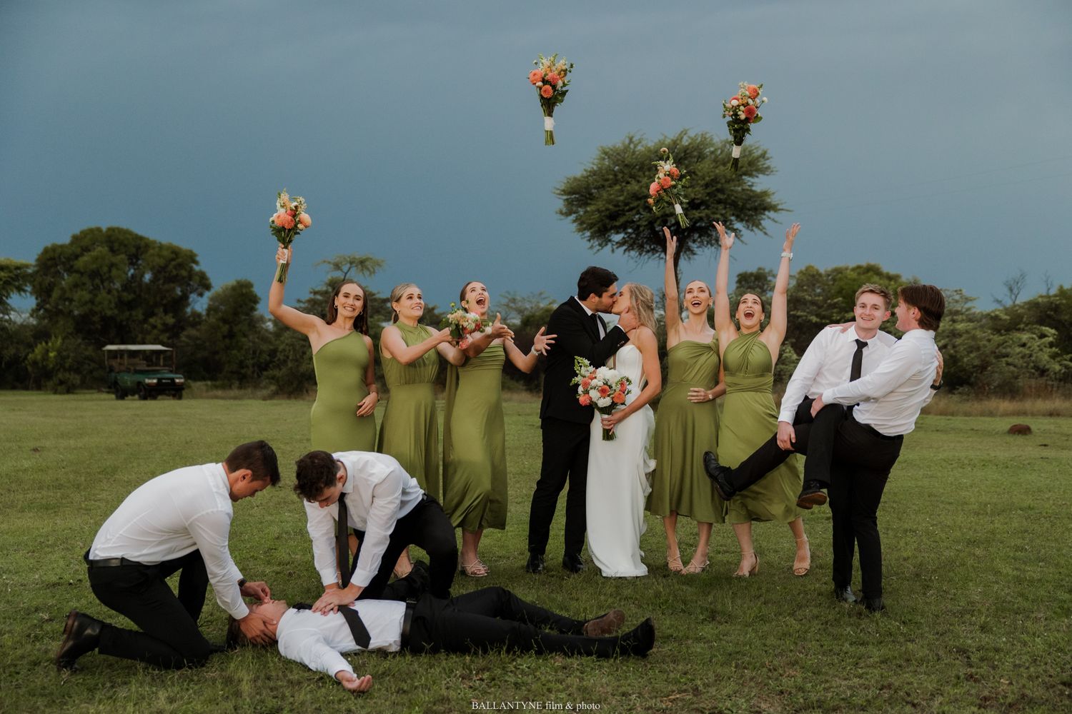 Wedding party celebrates outdoors, bridesmaids in olive gowns toss bouquets as bride and groom kiss on lush green lawn.