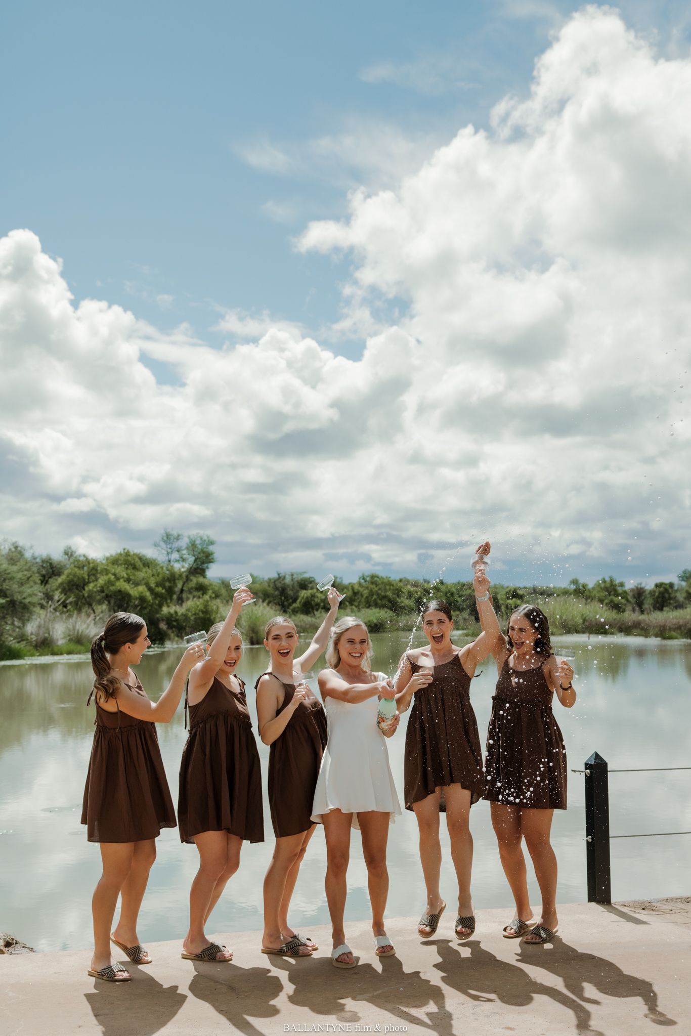 Six women in brown and white dresses raise glasses outdoors by a lake under a cloudy blue sky.