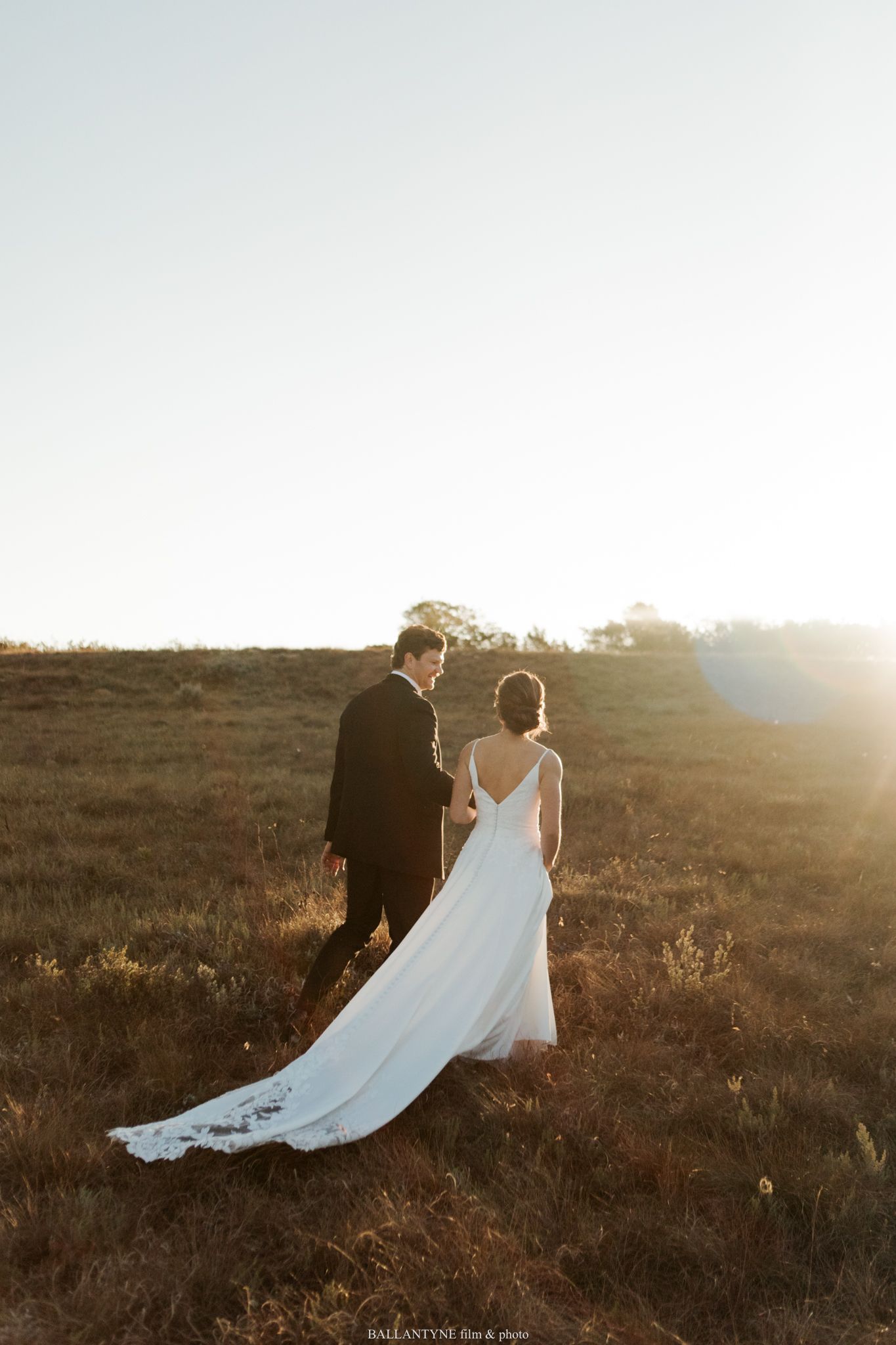 Wedding couple walking hand in hand across a field at sunset with dramatic lighting.