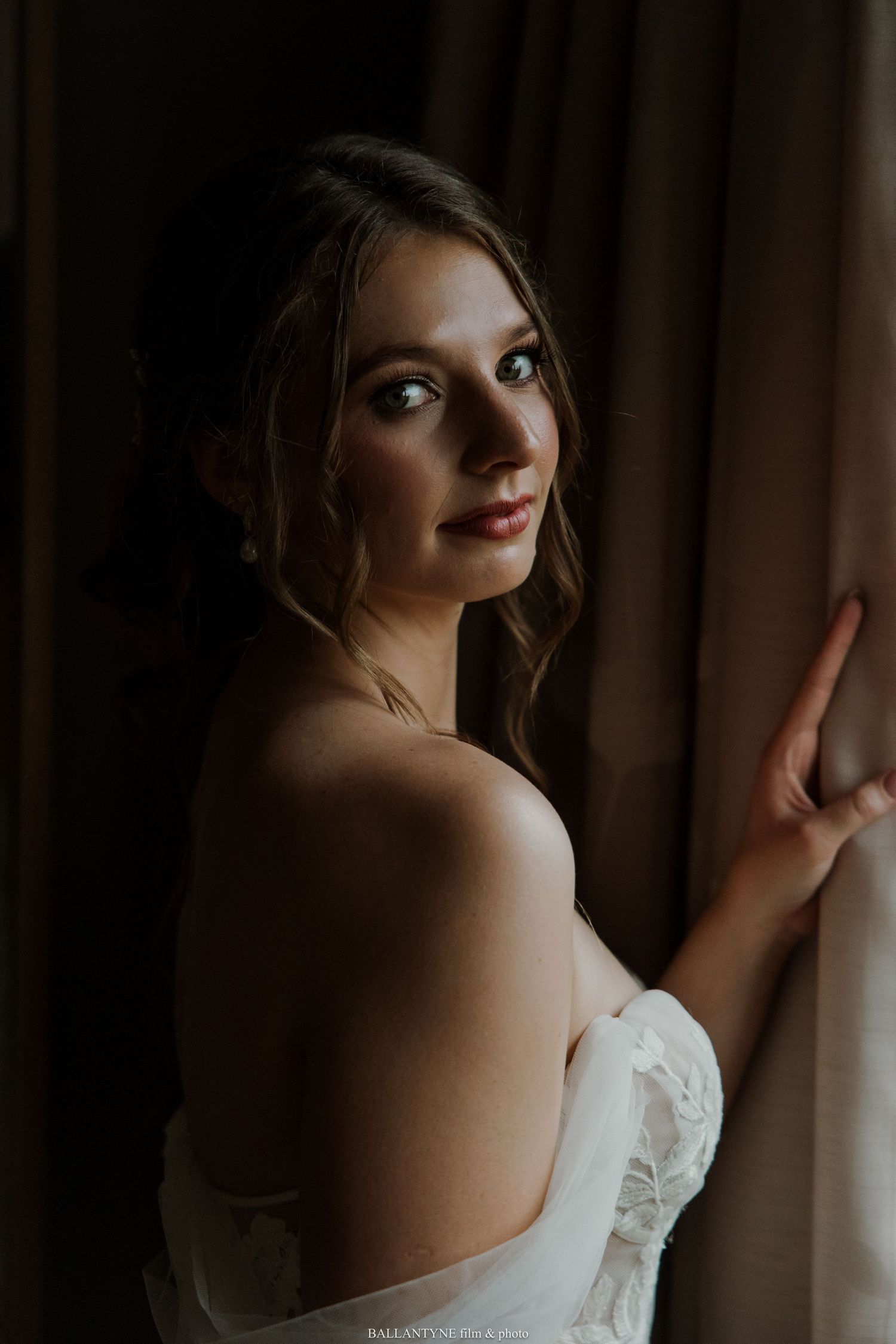 A romantic bridal portrait in a white lace gown near a window with dramatic lighting.
