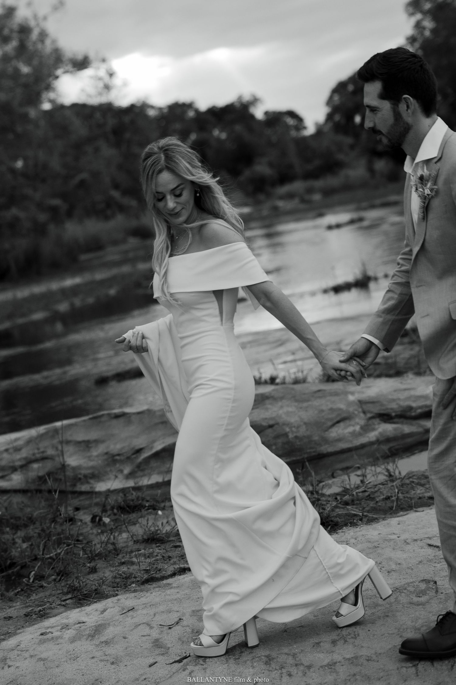 Black and white photo of bride walking carefully across rocky terrain in flowing dress.