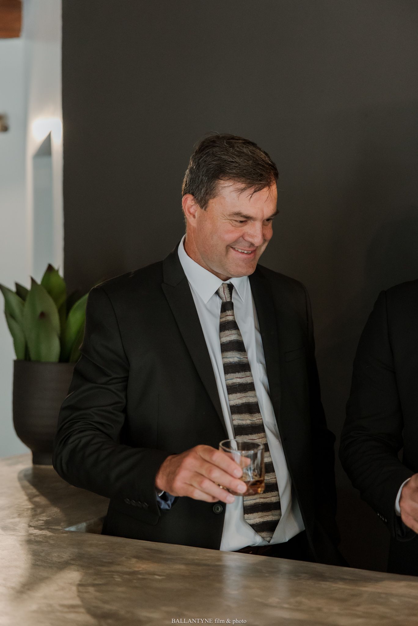 A smiling man in a black suit and striped tie holds a whiskey glass at a bar counter.
