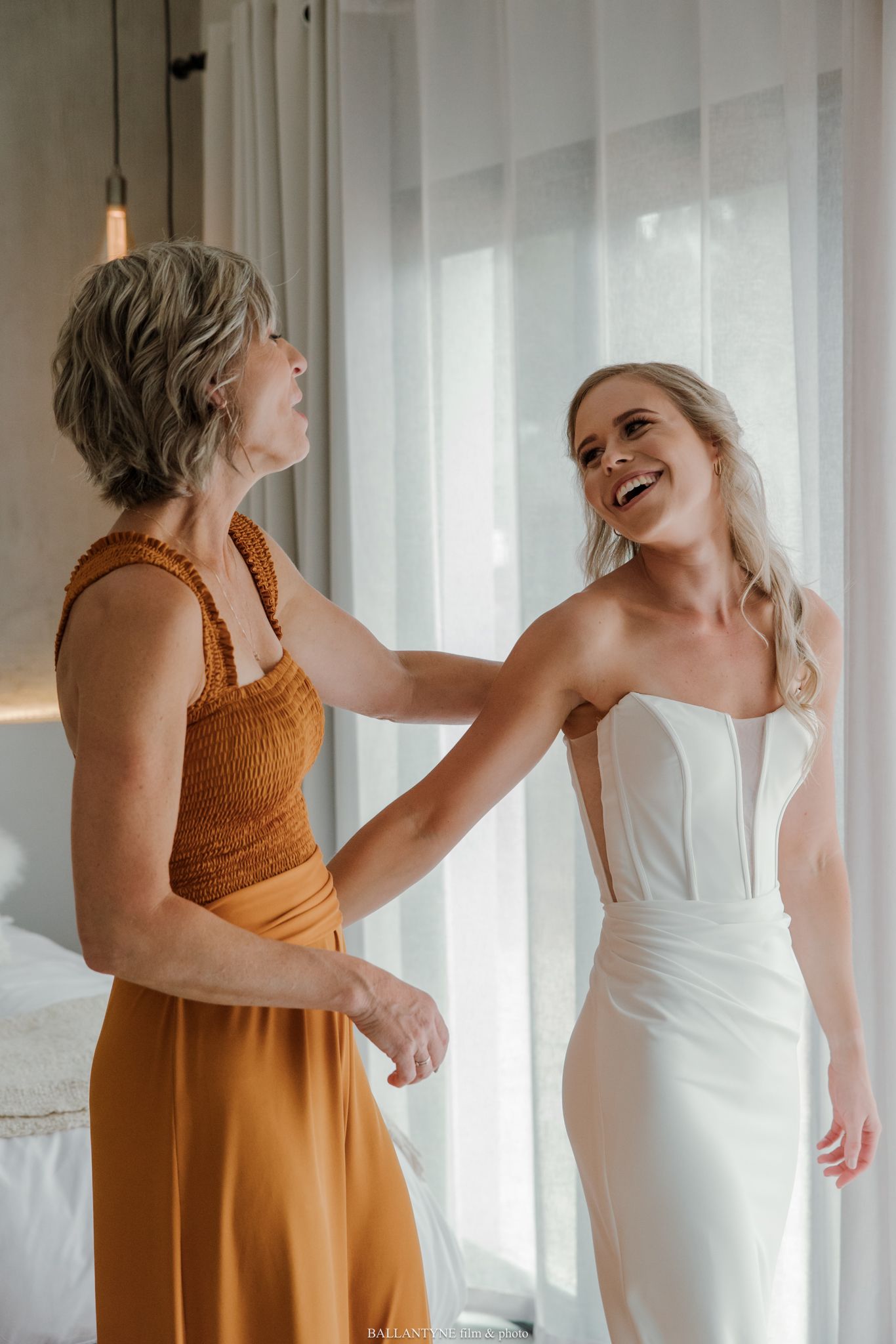 A bride in a white gown laughs joyfully as an older woman in orange helps her prepare for her wedding day.