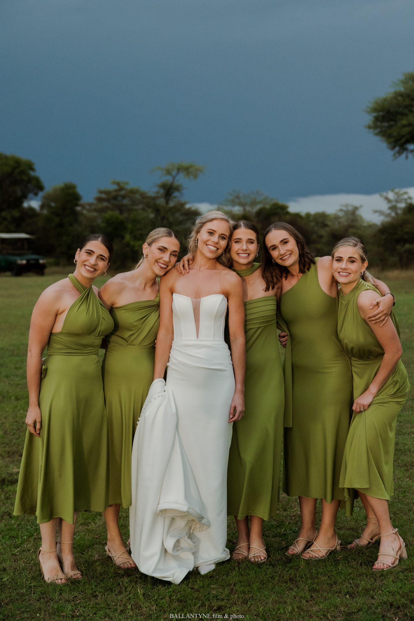Bride in white gown poses with five bridesmaids in olive green dresses on grass with stormy sky backdrop.