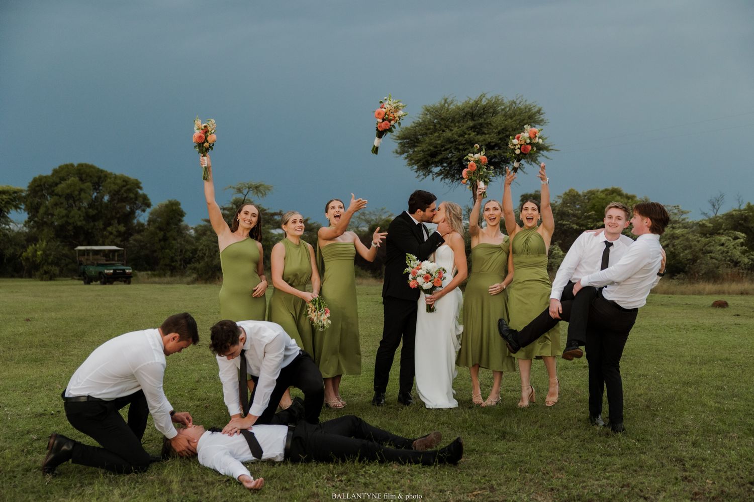 Wedding party celebrates outdoors, bridesmaids in green dresses raise bouquets as couple kisses while groomsmen pose playfully.