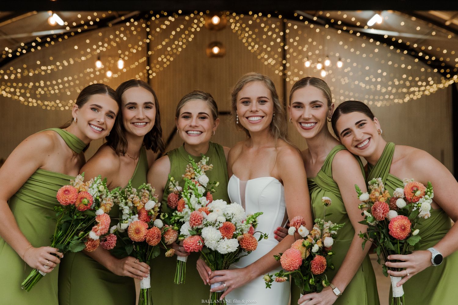 Bride in white gown with five bridesmaids in green dresses holding coral and white bouquets at a wedding reception.