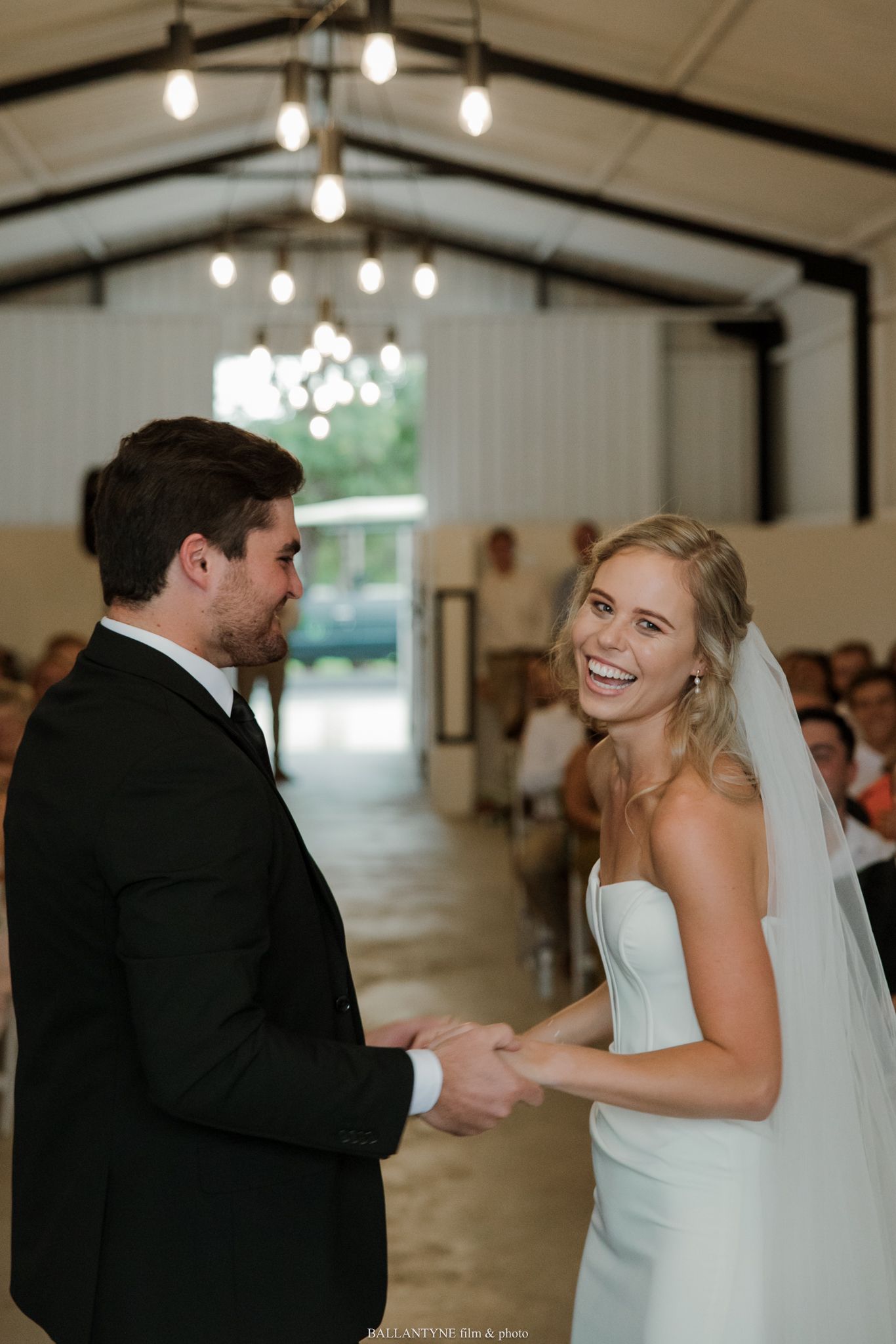 Bride and groom holding hands and smiling during wedding ceremony in elegant barn venue with Edison bulb lighting.