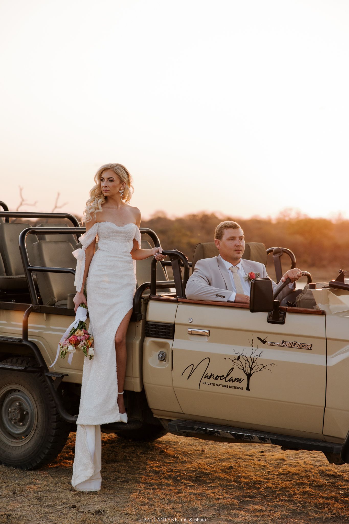 A wedding party poses near a vintage safari vehicle during golden hour.