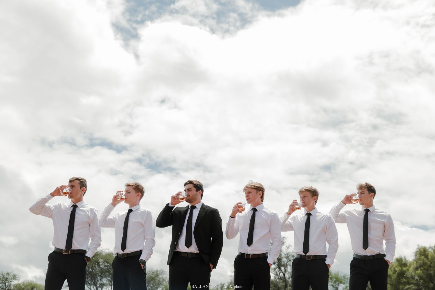 Six groomsmen in white shirts and black ties raise glasses for a toast outdoors under a cloudy sky.