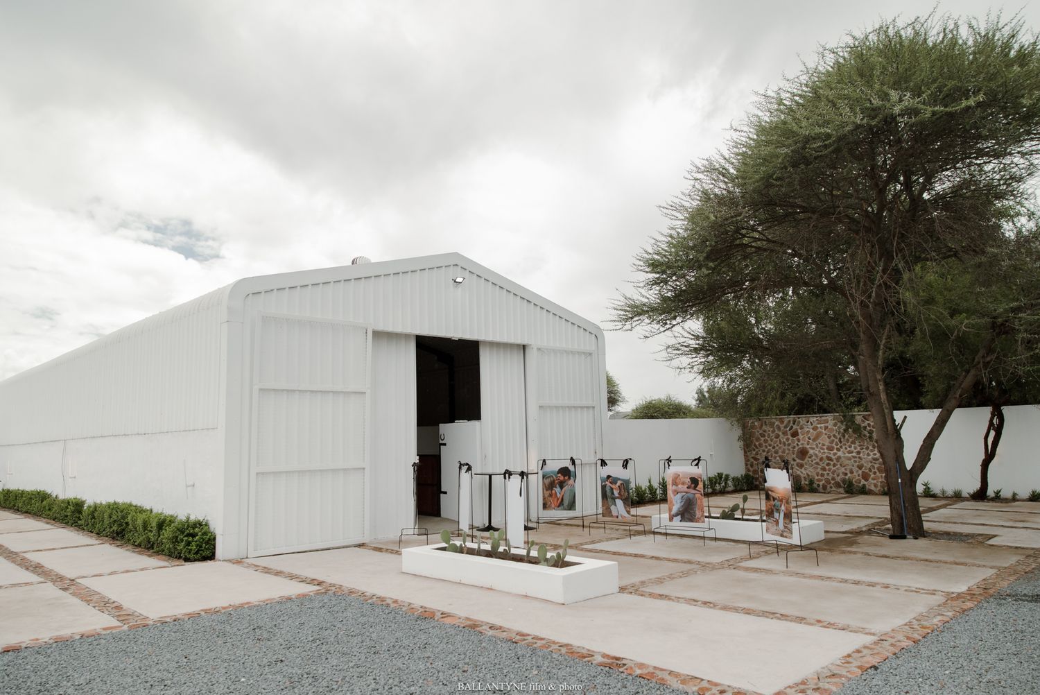 White metal barn building with open doors, outdoor seating area, gravel path, and a large tree under cloudy skies.