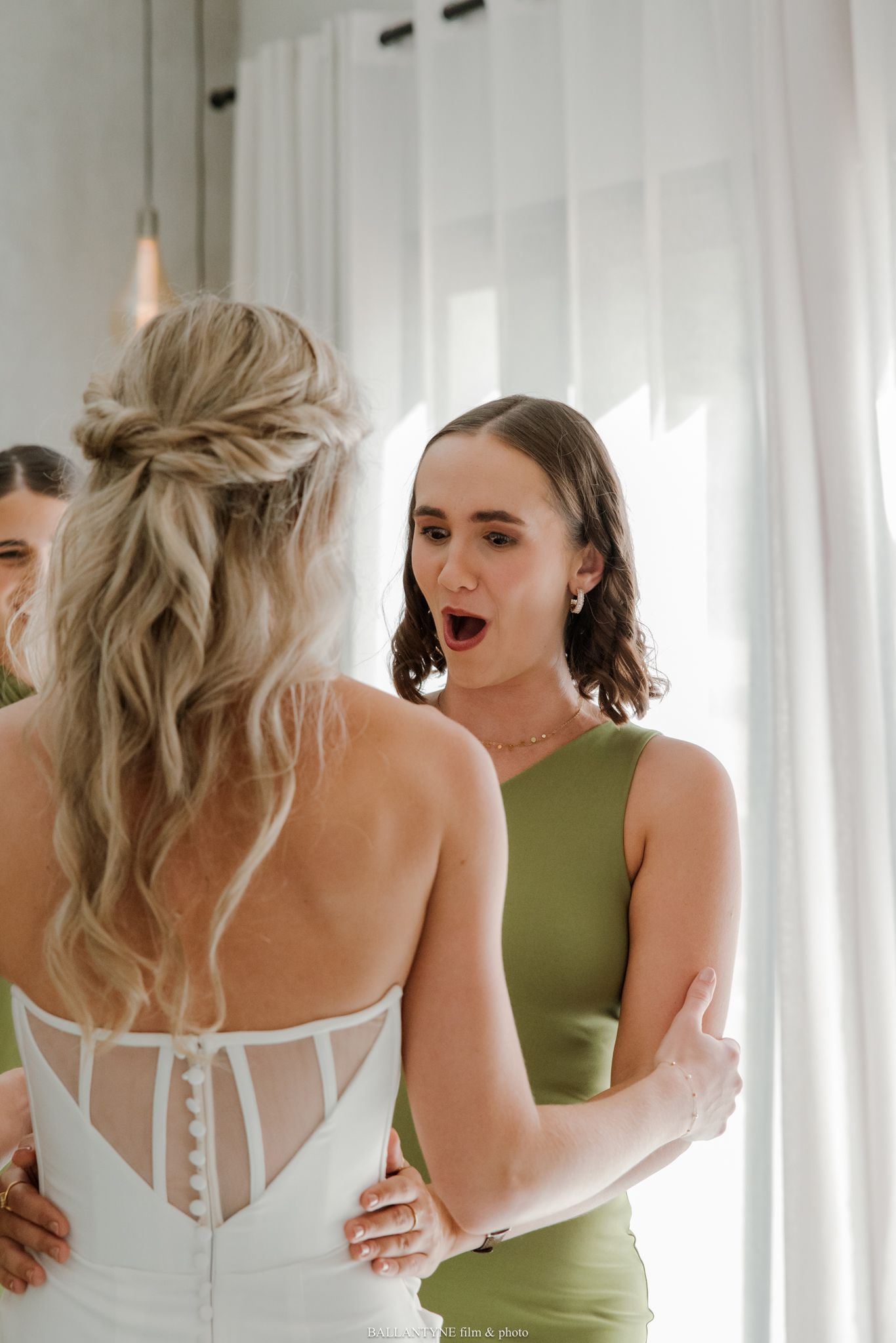 Bridesmaid in green dress reacts with awe seeing bride in white corset wedding gown during bridal preparations.