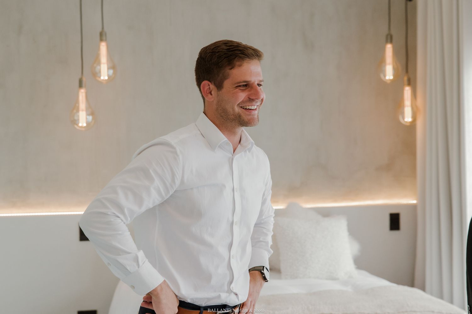Smiling man in white dress shirt standing confidently in a modern hotel room with pendant Edison bulb lighting.