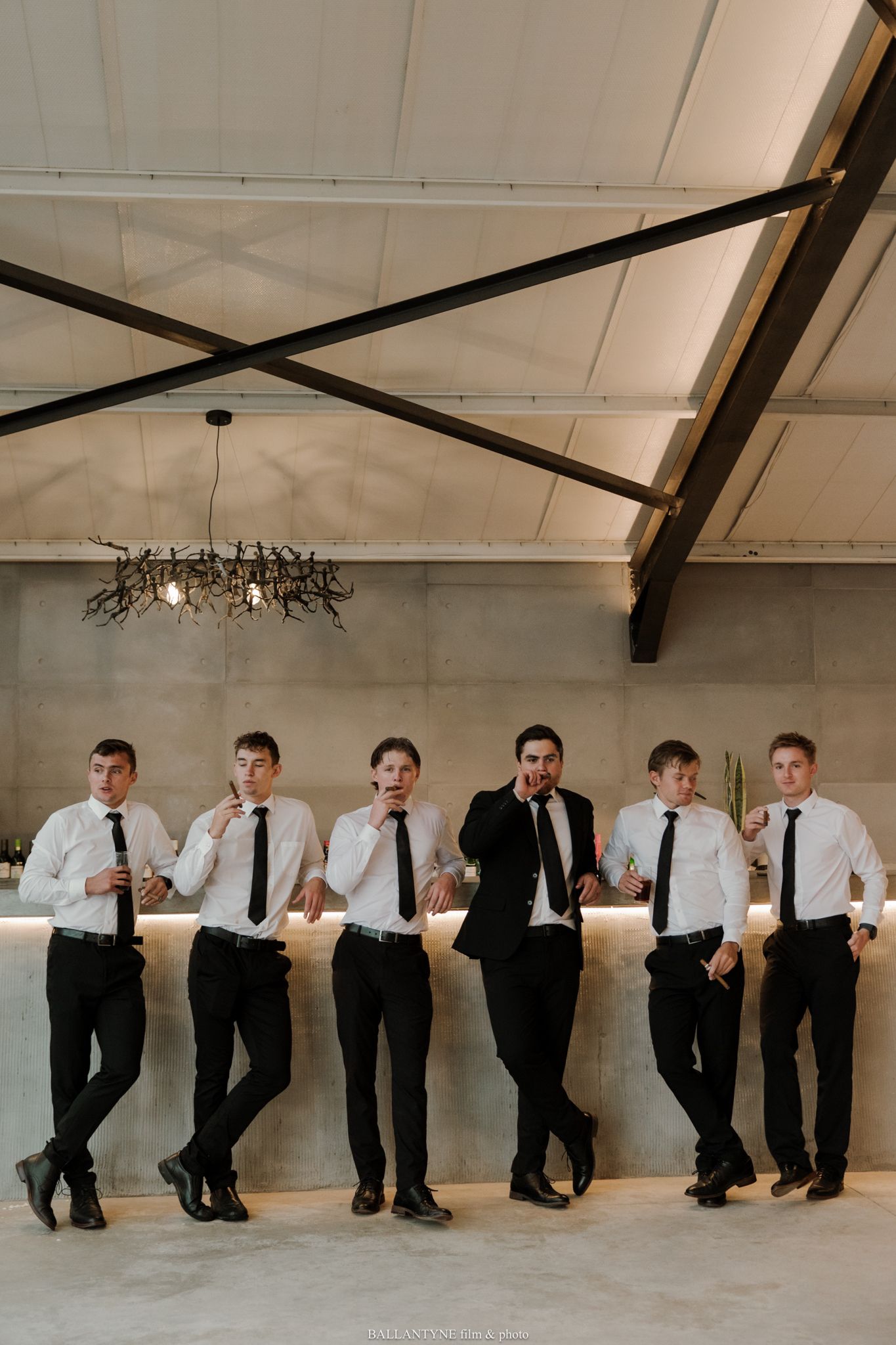 Groom in black suit stands with six groomsmen in white shirts and black ties, leaning against a bar at a wedding venue.
